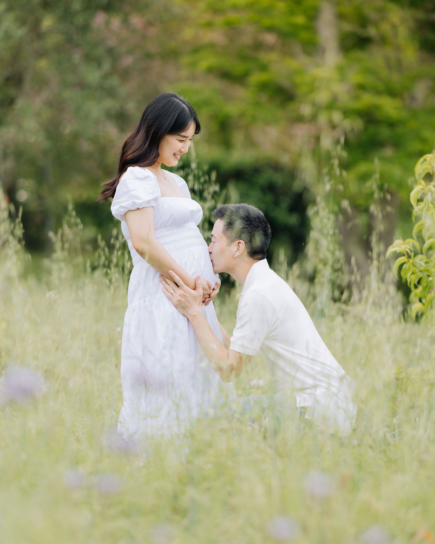 Man kneeling to kiss pregnant woman's belly in lush green meadow, both wearing white Filoli Woodside maternity photoshoot.