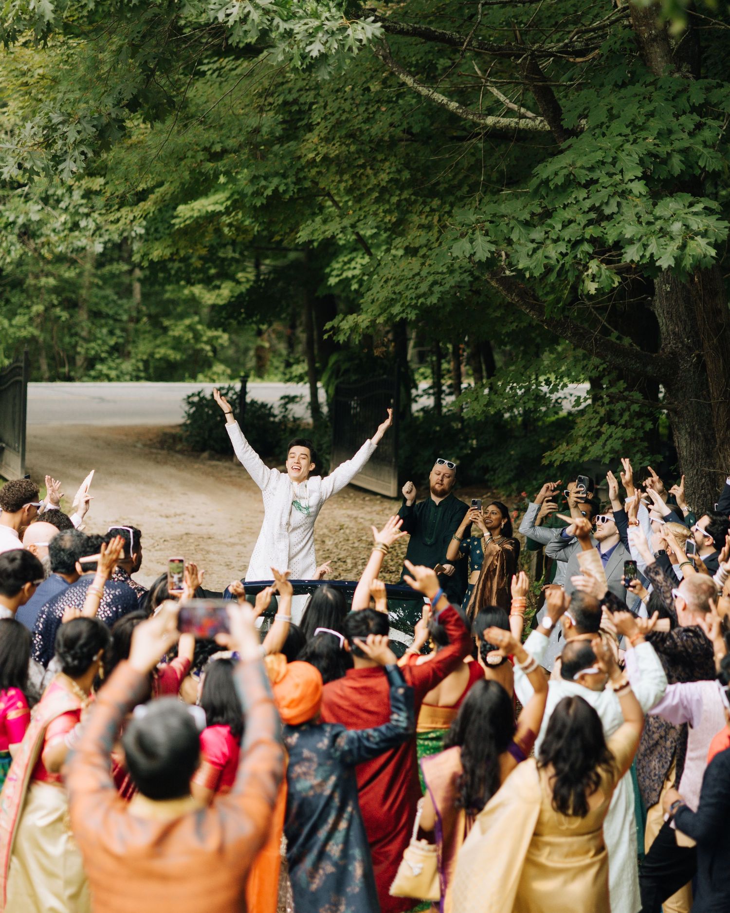 Groom celebrates with raised arms during vibrant outdoor Indian wedding baraat ceremony surrounded by joyful guests.