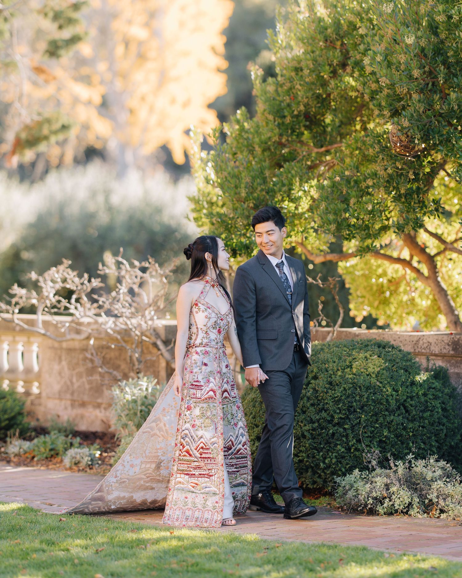 Couple holding hands in garden, woman in detailed embroidered gown glancing back at partner during Filoli engagement photoshoot.