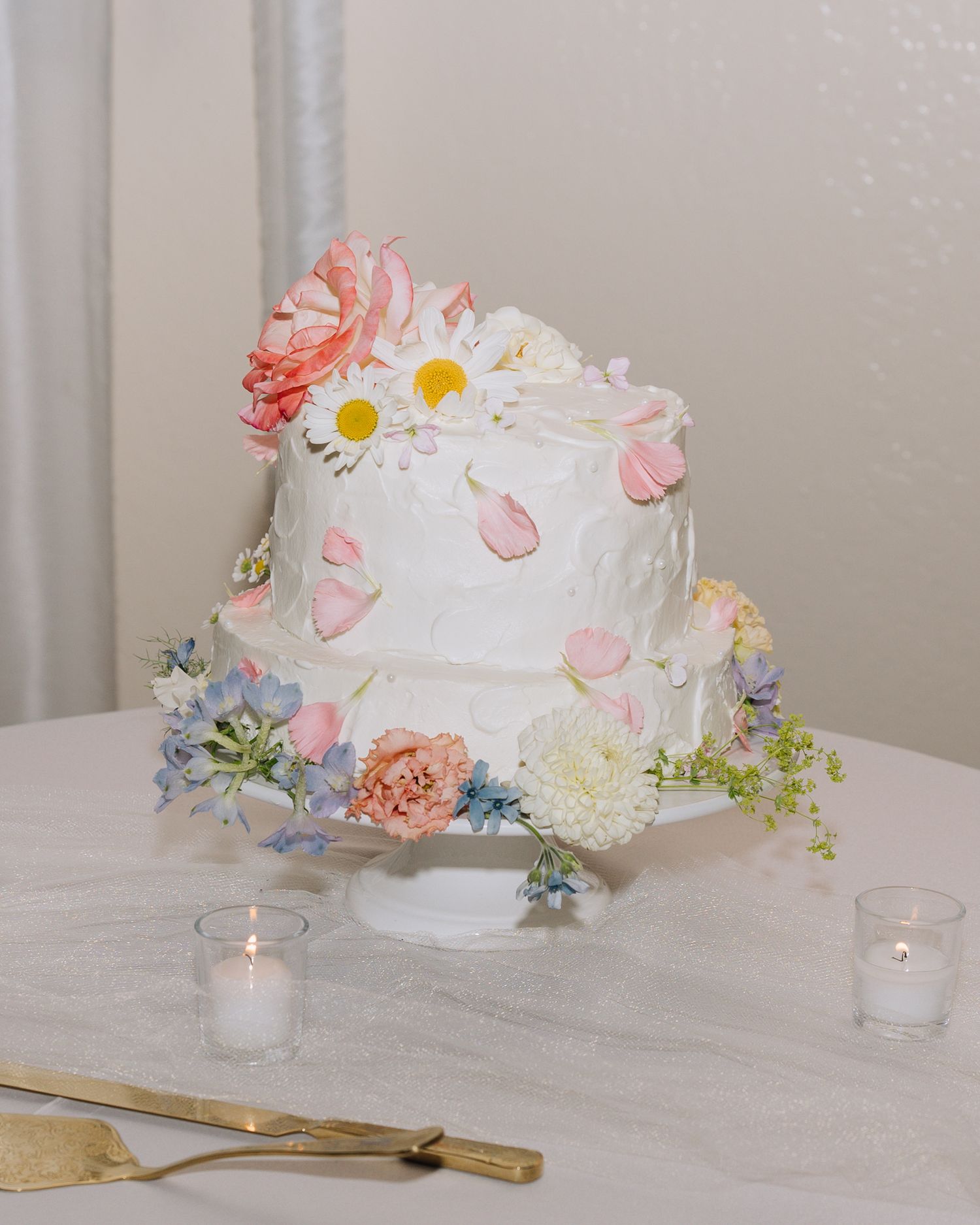 Elegant two-tier white wedding cake decorated with colorful fresh flowers including daisies, pink roses, and blue blooms.