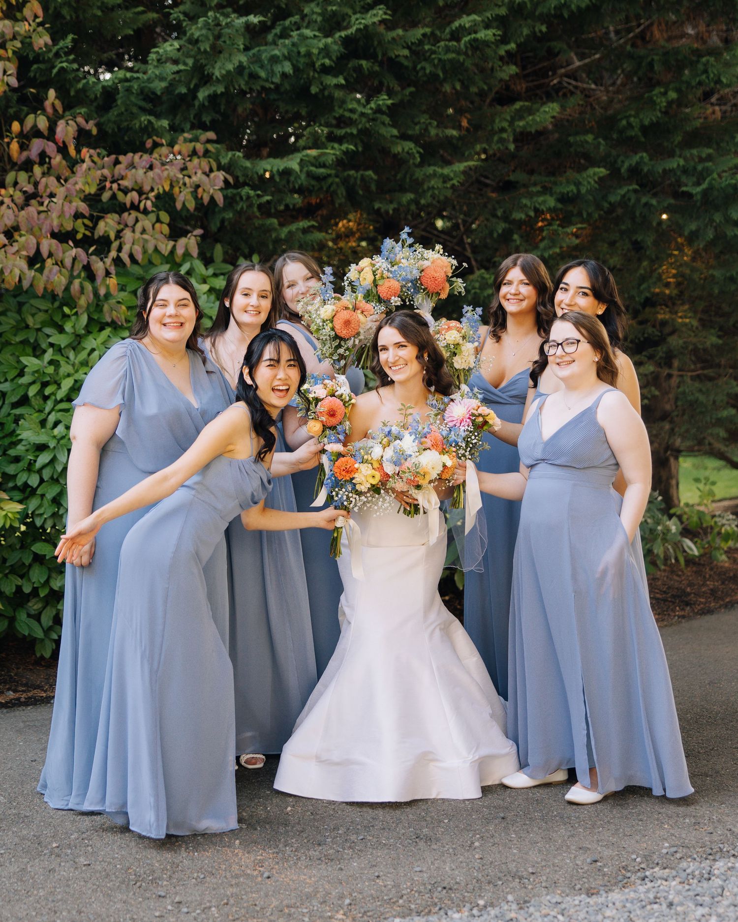 Bride in white gown poses with six bridesmaids in dusty blue dresses holding colorful bouquets outdoors. Wedding at the Hall at Fauntleroy