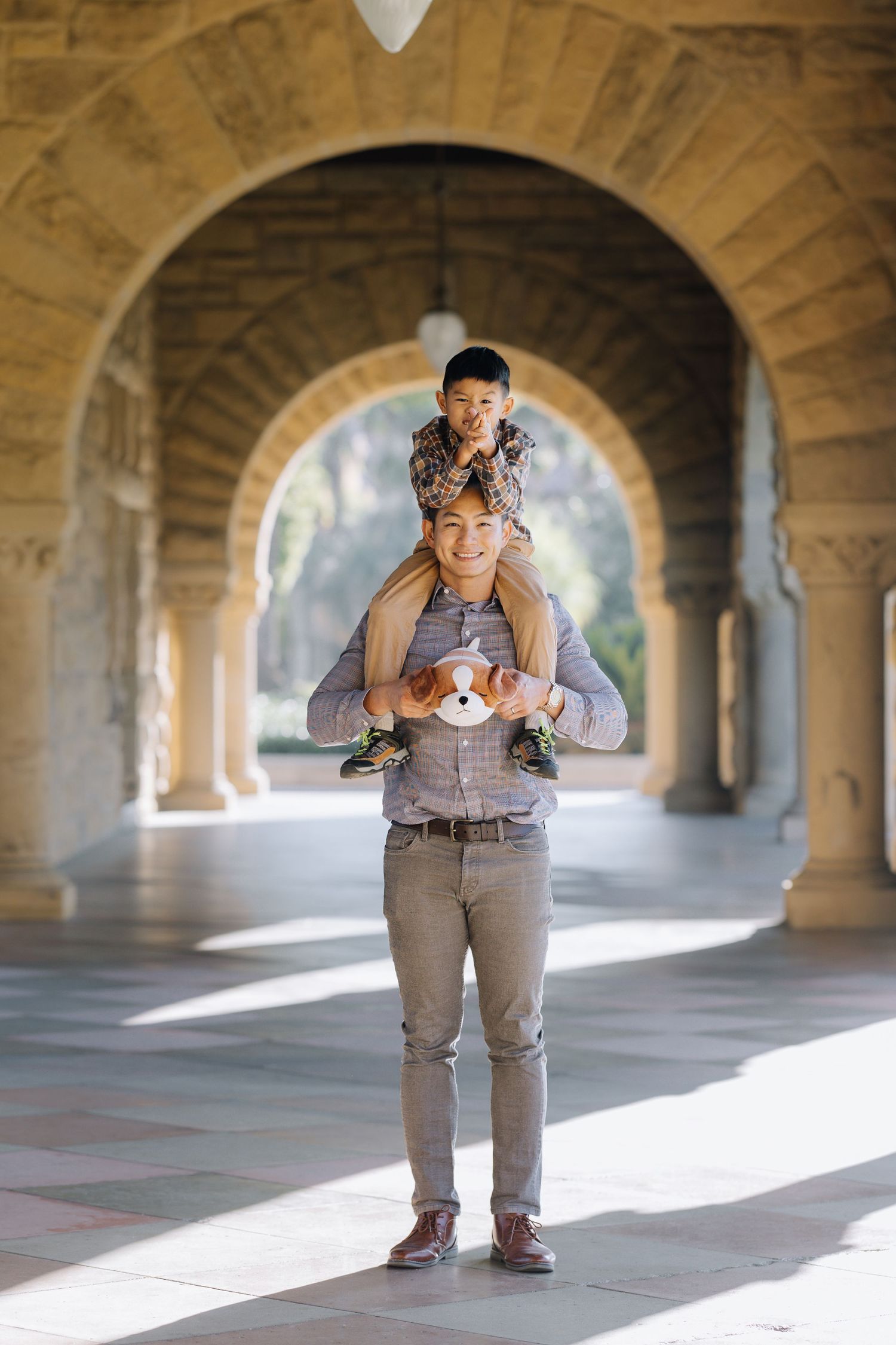 Smiling Asian dad carrying young son on shoulders under stone archway corridor at Stanford family photoshoot.