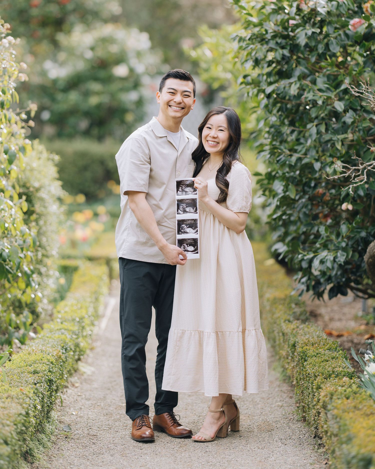 Smiling couple holding ultrasound photos on a garden pathway surrounded by manicured hedges and flowers at Filoli Woodside maternity session