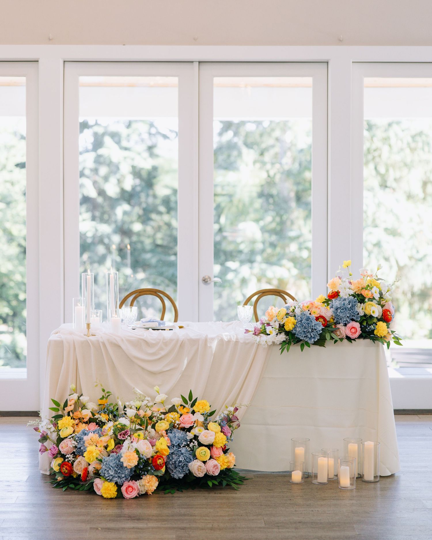 Sweetheart table and gift table adorned with lush colorful floral arrangements of blue, yellow, and pink flowers.