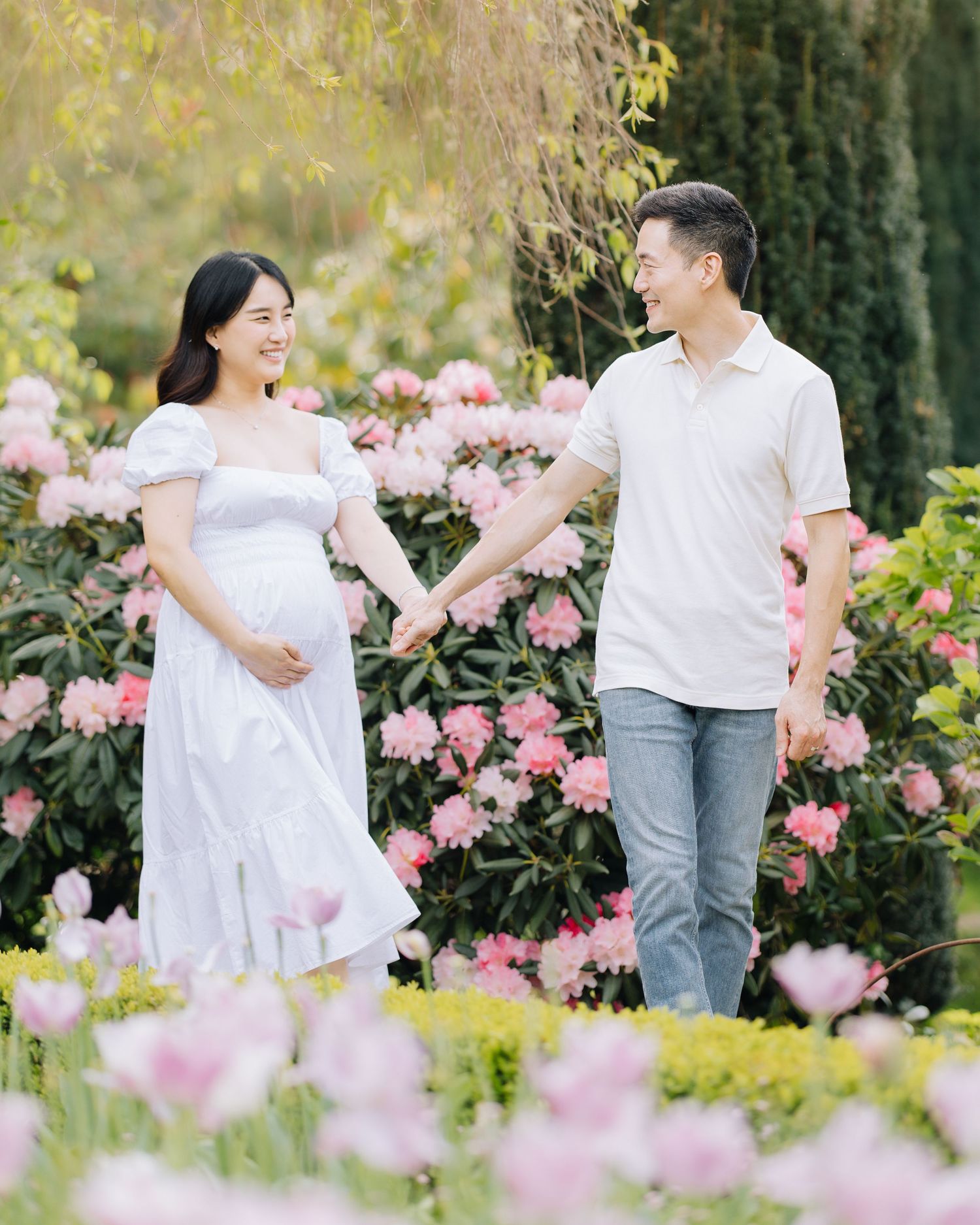 Pregnant woman in white dress holding hands with partner among pink spring flowers at Filoli Woodside maternity photoshoot.