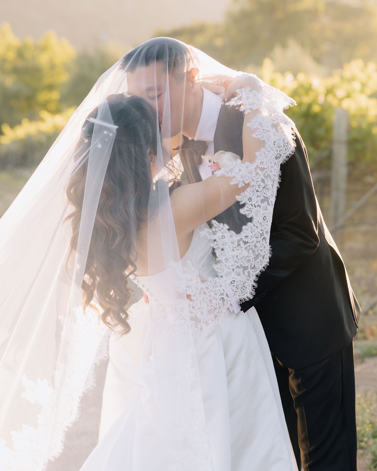 Bride and groom share a kiss under a lace-trimmed veil in a golden vineyard at sunset at Bernardus Lodge in Carmel Valley,