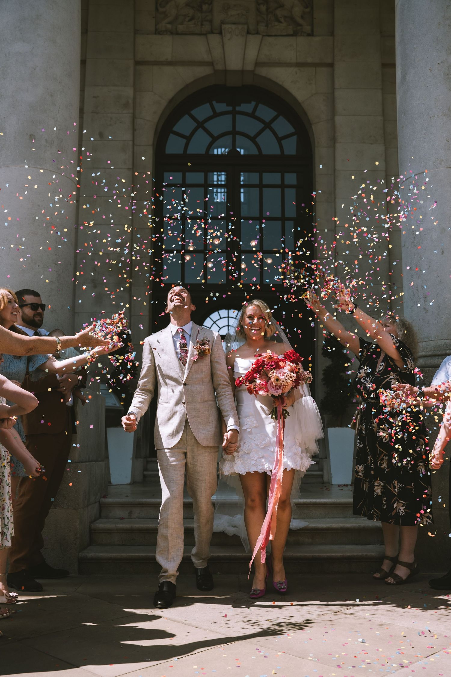 Couple walks down sunlit steps as guests toss colorful confetti outside elegant stone building in Lancaster at Ashton Memorial