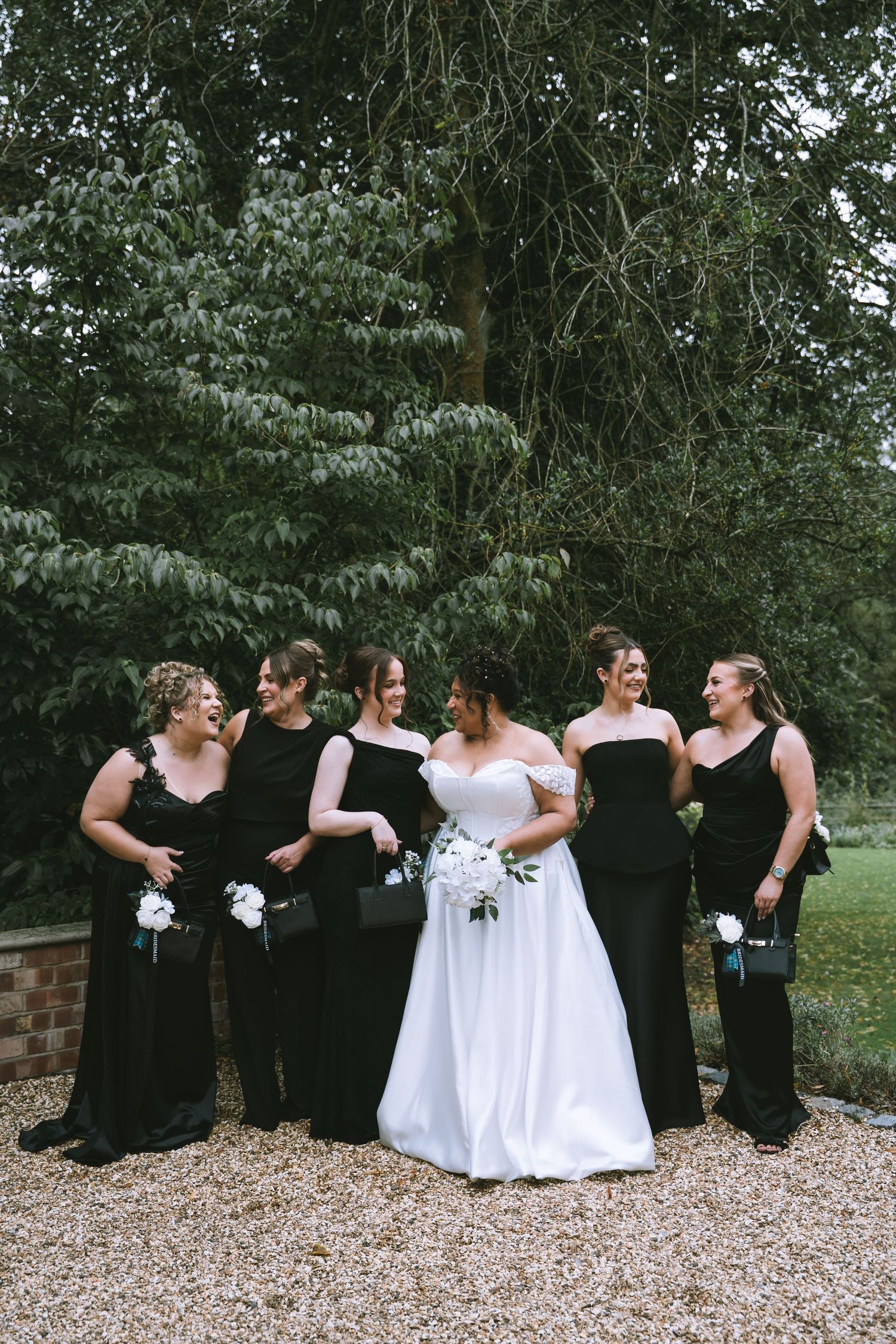 Bride in white off-shoulder gown poses with five bridesmaids in black dresses outdoors.