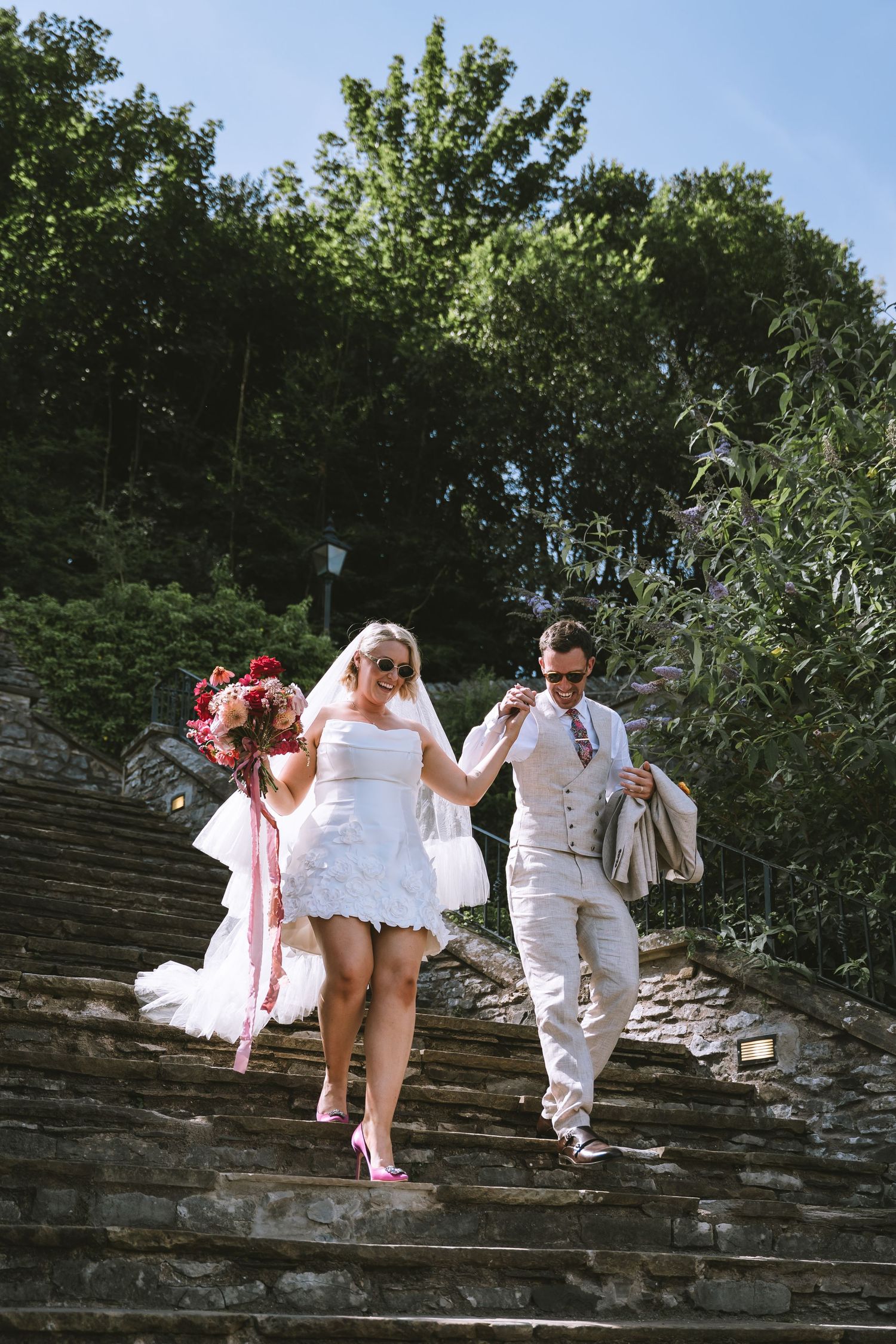 Bride and groom in sunglasses walk down stone steps holding hands on a sunny wedding day in The Lake District