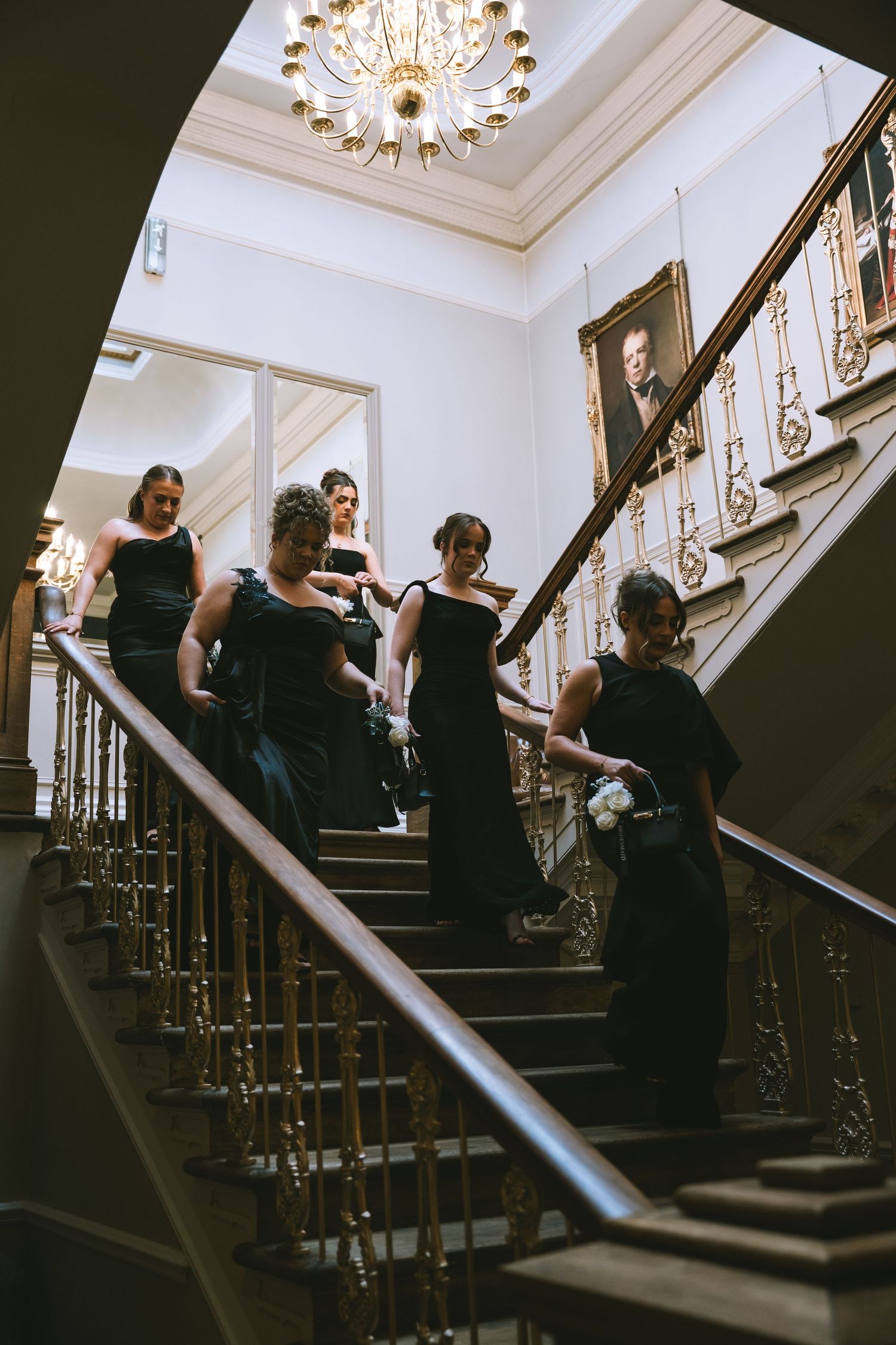 Bridesmaids in elegant black gowns descend grand staircase beneath ornate chandelier in luxury venue.