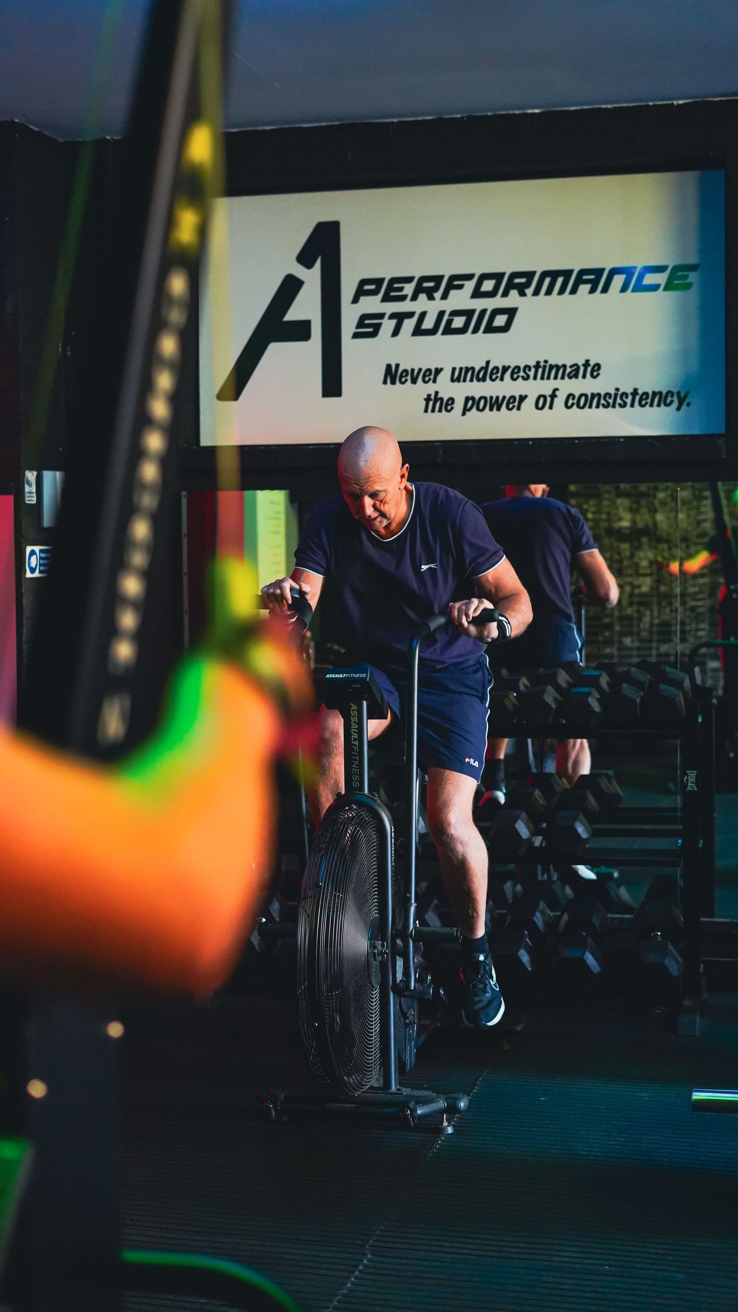 Bald man cycling on air bike at A1 Performance Studio with motivational consistency quote on banner.