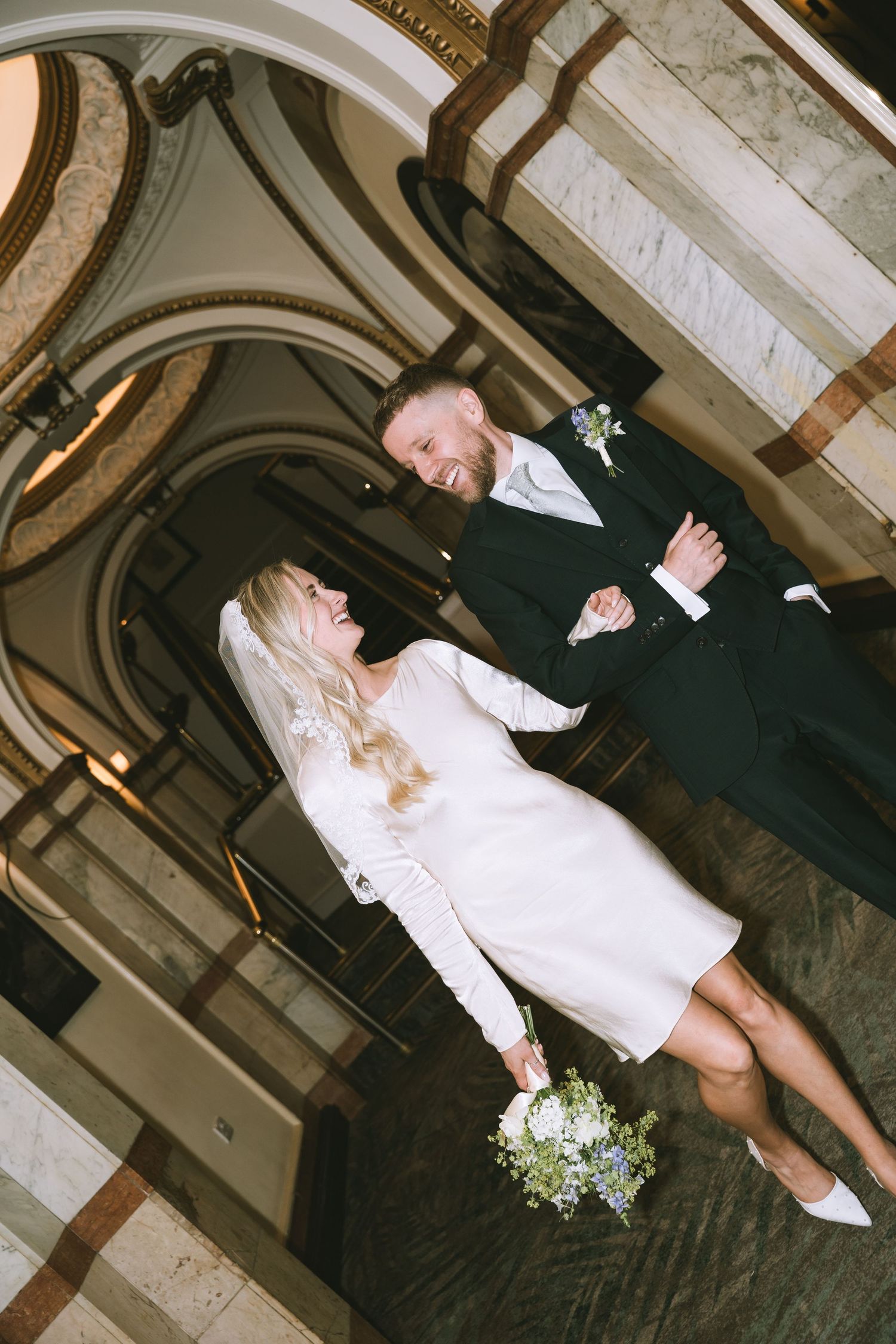 Bride in short white long-sleeve dress and groom in dark suit laugh together inside grand marble building in Manchester