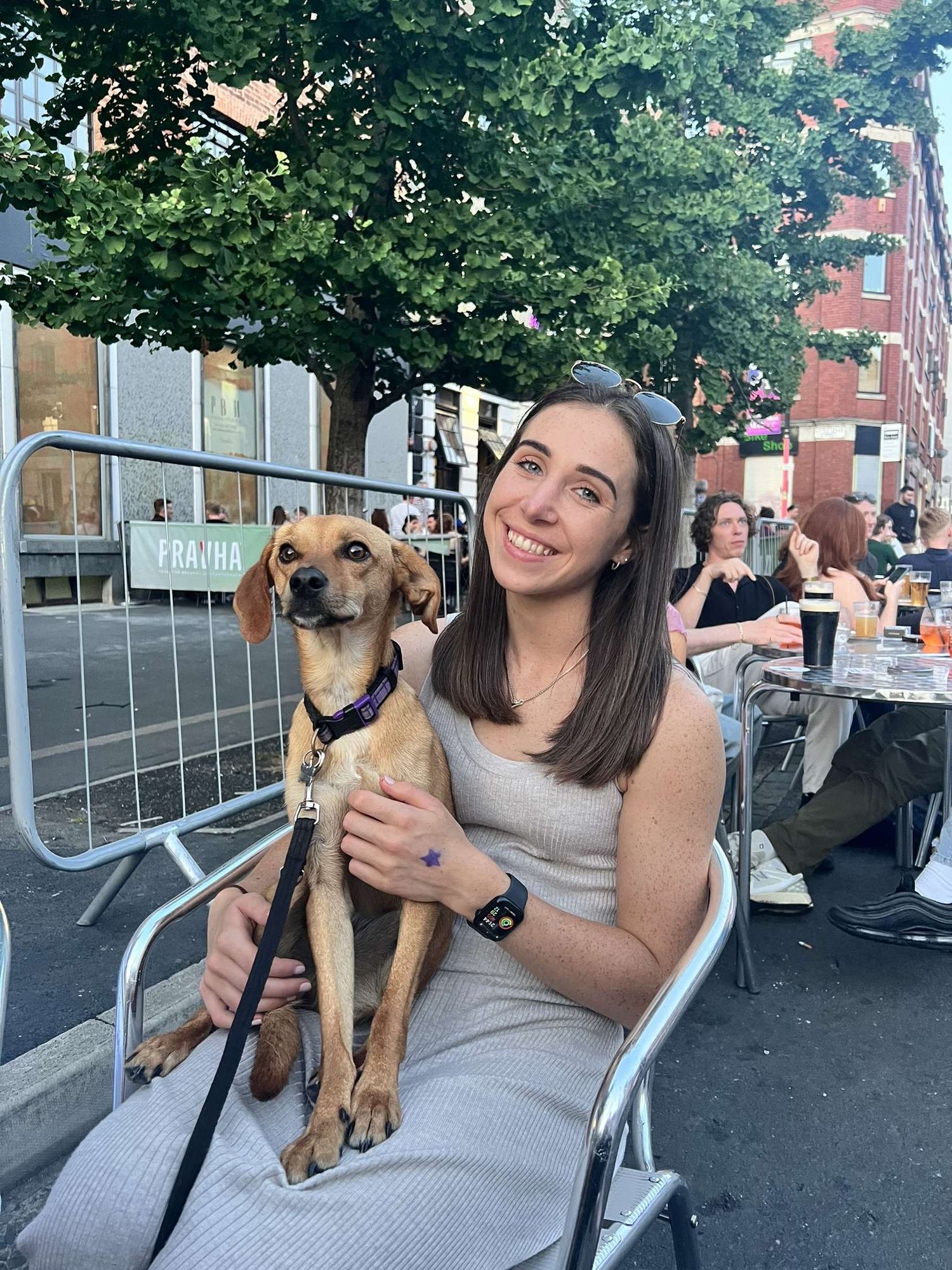 A smiling woman holds a small brown dog on a leash at an outdoor cafe patio with trees and city street in background.
