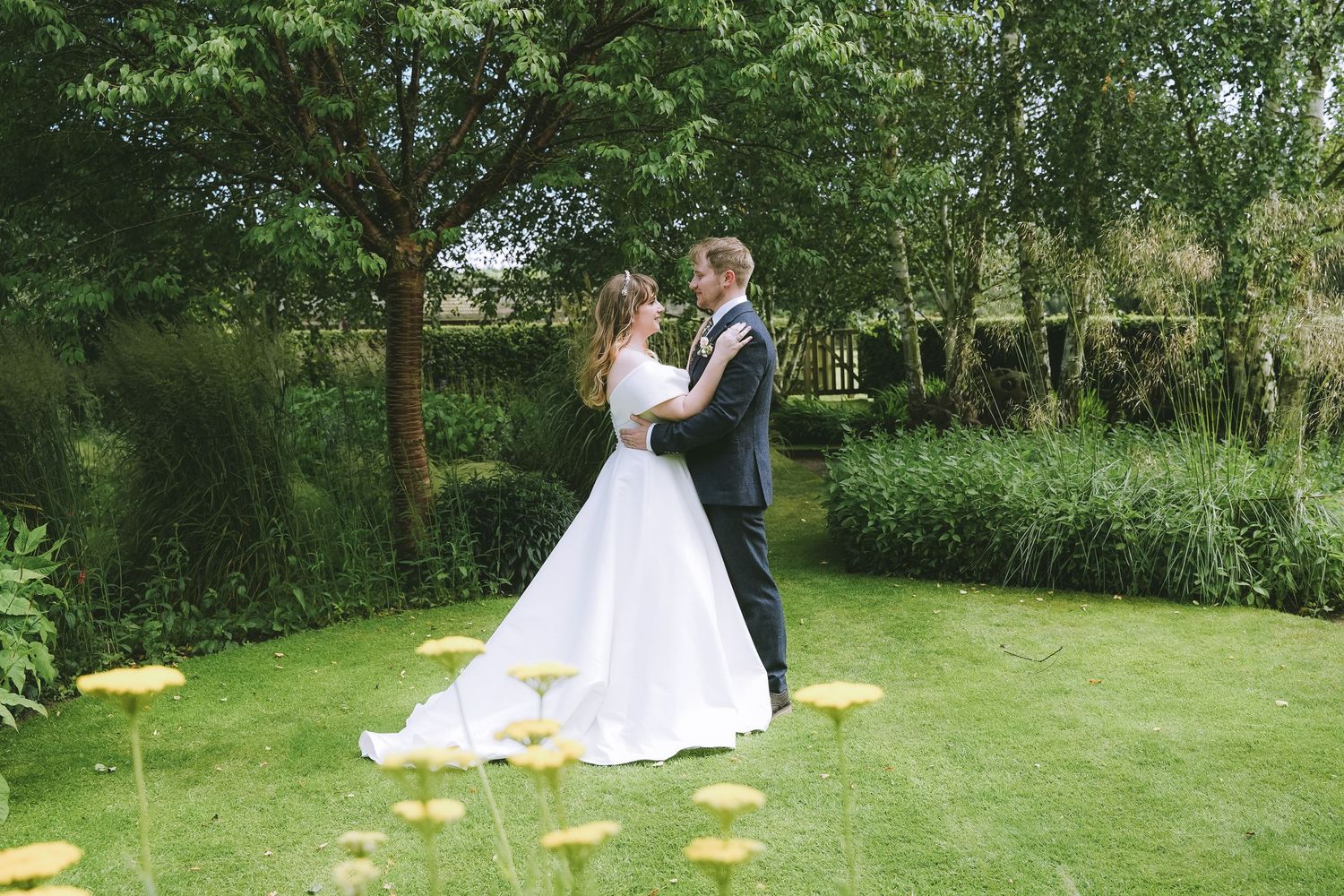 Bride and groom embrace in lush green garden with yellow flowers, sharing a romantic moment on their wedding day.
