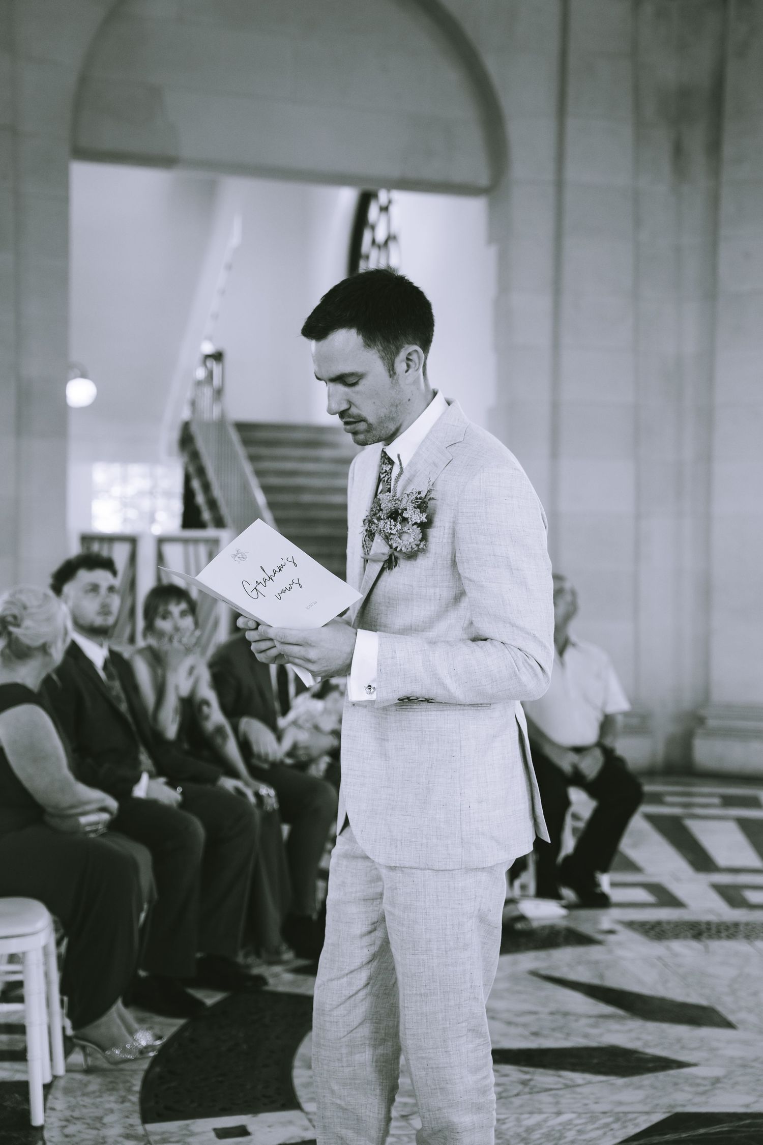 Groom in a light linen suit reads from a wedding program during the ceremony in a grand venue in Lancaster at Ashton Memorial