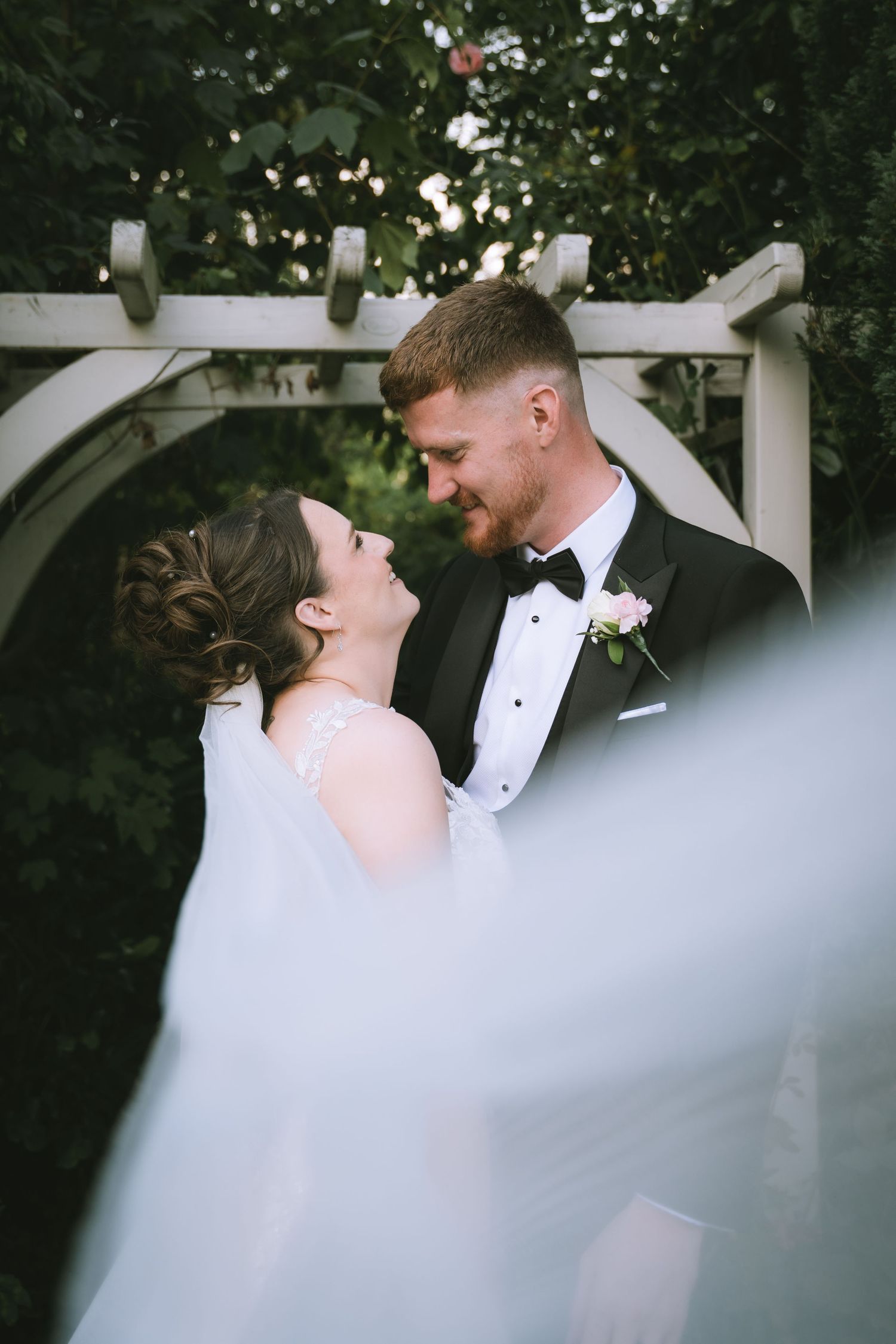 Bride and groom share a romantic moment under a white garden arbor, smiling at each other on their wedding day.