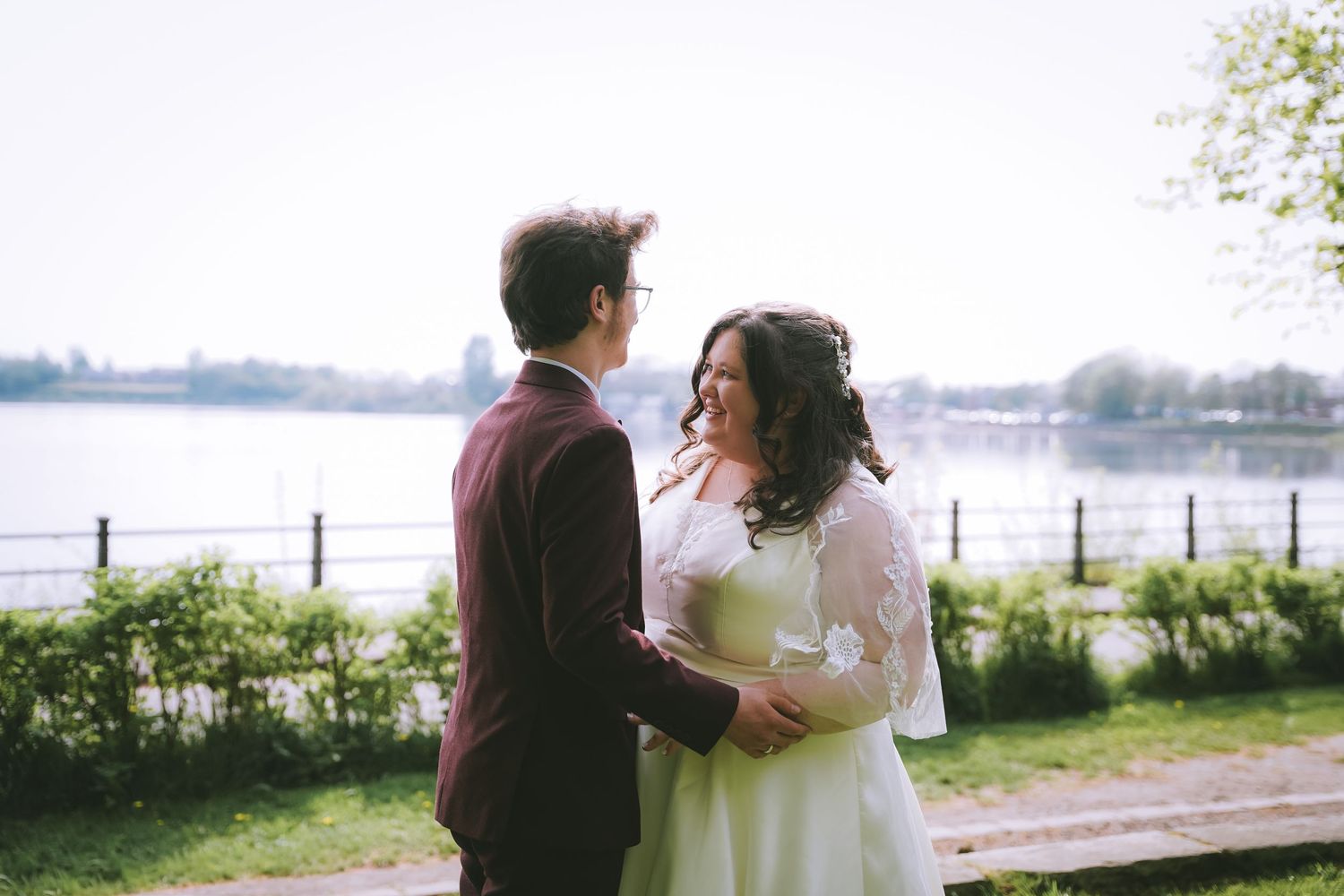 Newlyweds gaze lovingly at each other in park with serene lake backdrop on spring day.