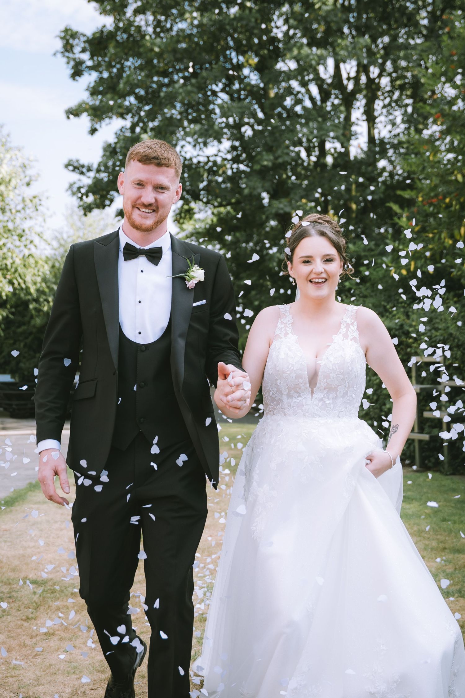Joyful bride and groom walk through confetti shower outdoors, groom in black tuxedo, bride in lace gown.