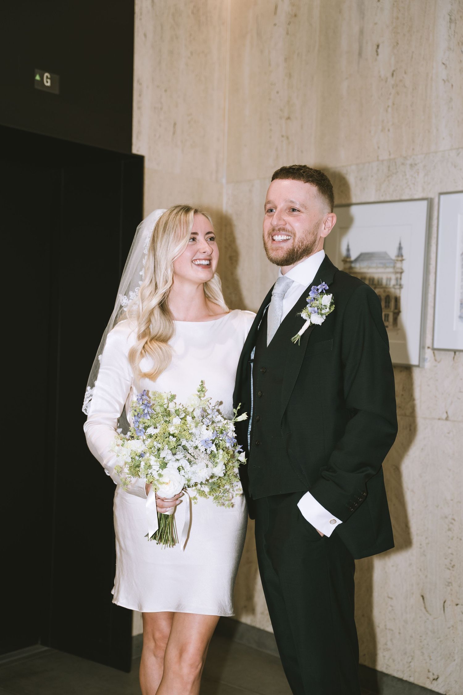 Smiling bride in short white dress holding wildflower bouquet stands with groom in black suit indoors.