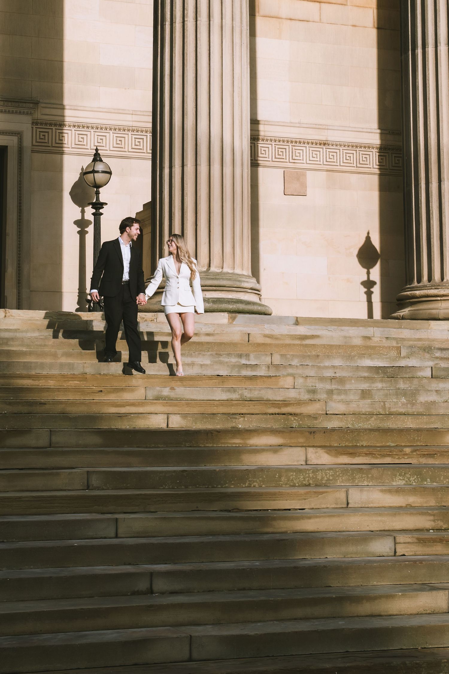 Bride and groom walk down grand stone steps hand in hand outside a classical columned building in Liverpool at St Georges Hall