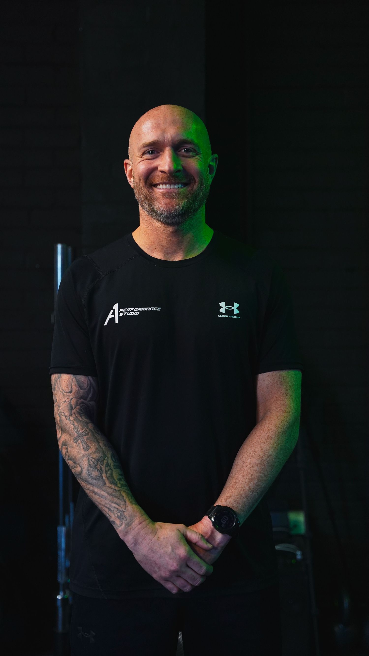 Smiling bald man with tattoos wearing black A1 Performance Studio and Under Armour t-shirt poses confidently.