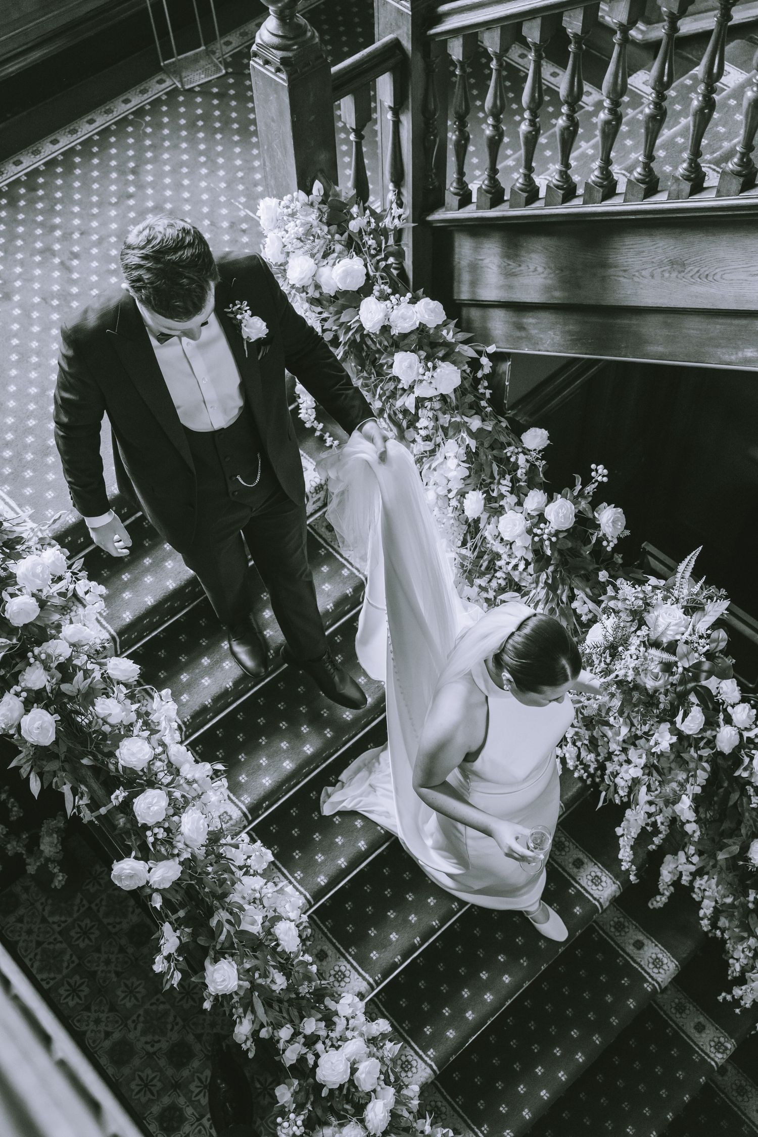 Aerial black and white shot of bride and groom descending floral-lined staircase in elegant wedding attire at Walton Hall & Gardens
