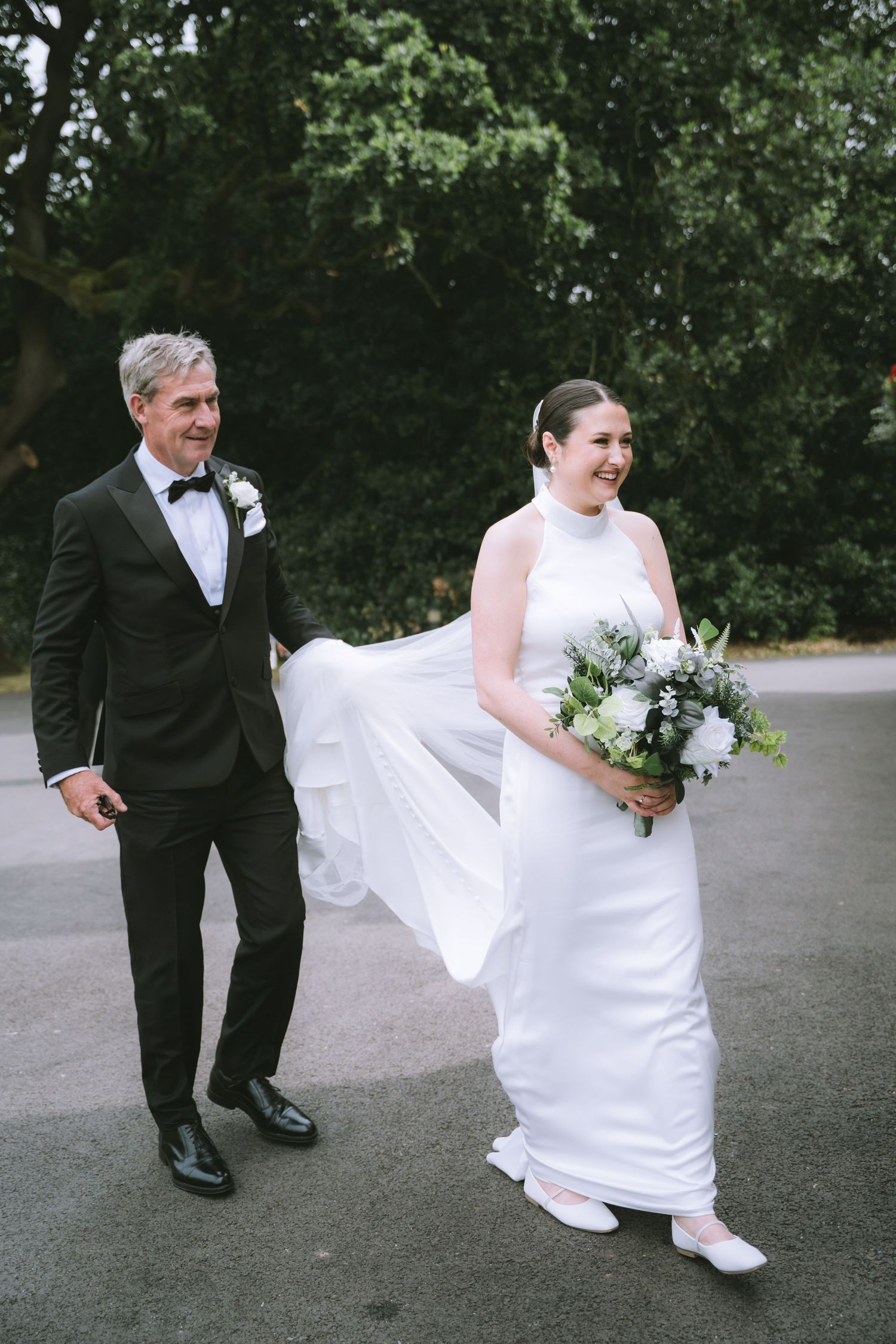 Smiling bride in a sleek white gown carries a white bouquet while walking with a man in a tuxedo at Walton Hall & Gardens
