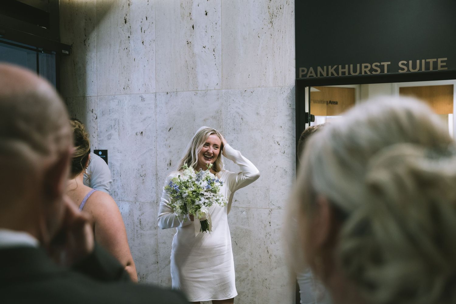 Smiling bride in short white dress holds bouquet of wildflowers outside the Pankhurst Suite wedding venue.