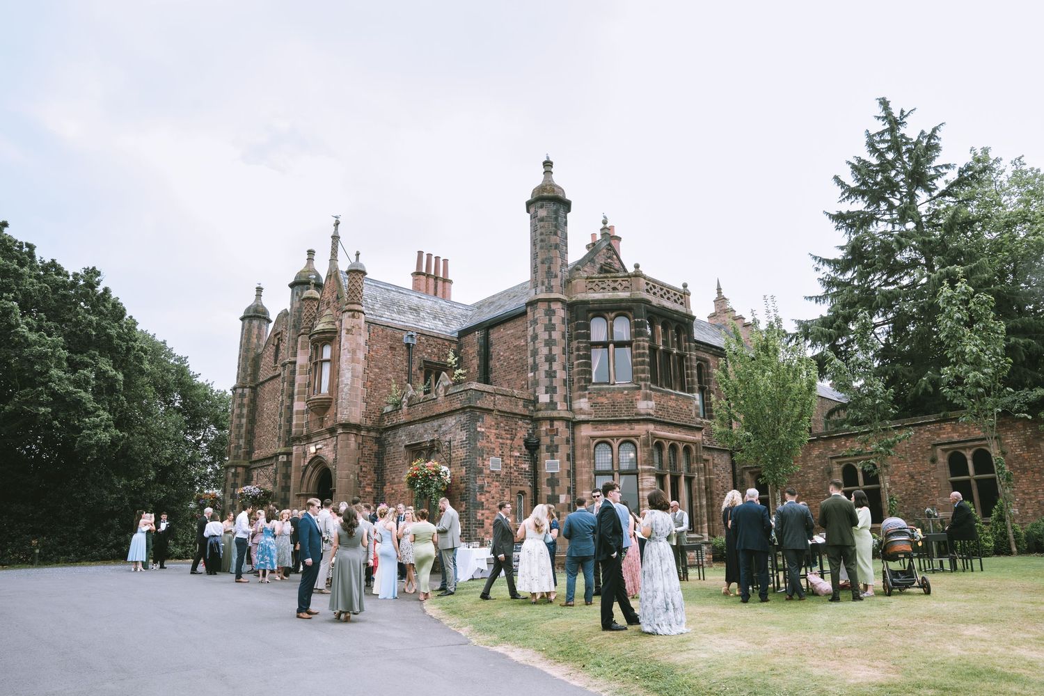 Wedding guests mingle outside a grand Victorian red brick manor house on a cloudy day at Walton Hall & Gardens in Warrington