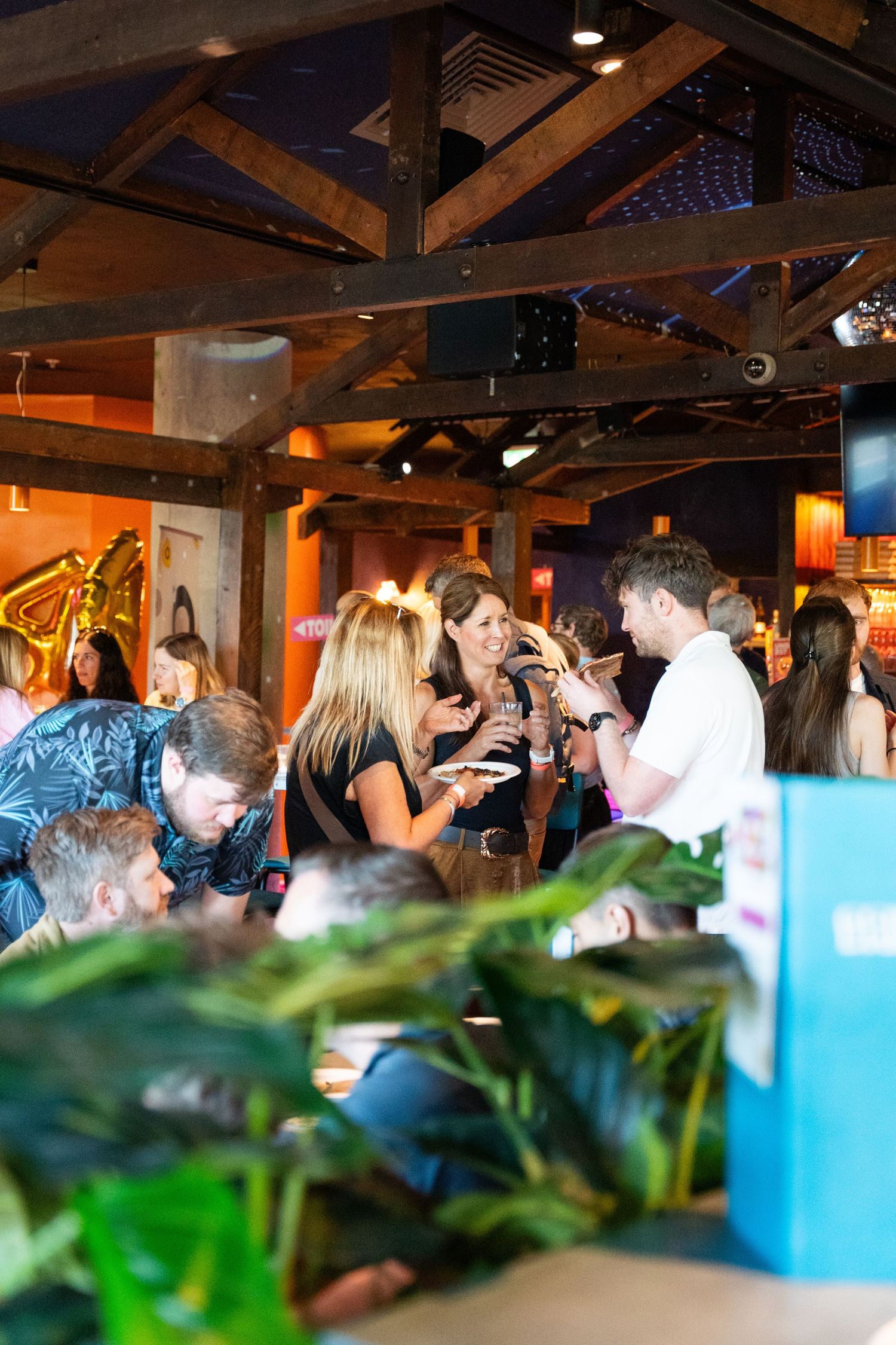 Guests mingling and enjoying drinks at a lively indoor event in a rustic venue with wooden beam ceiling.