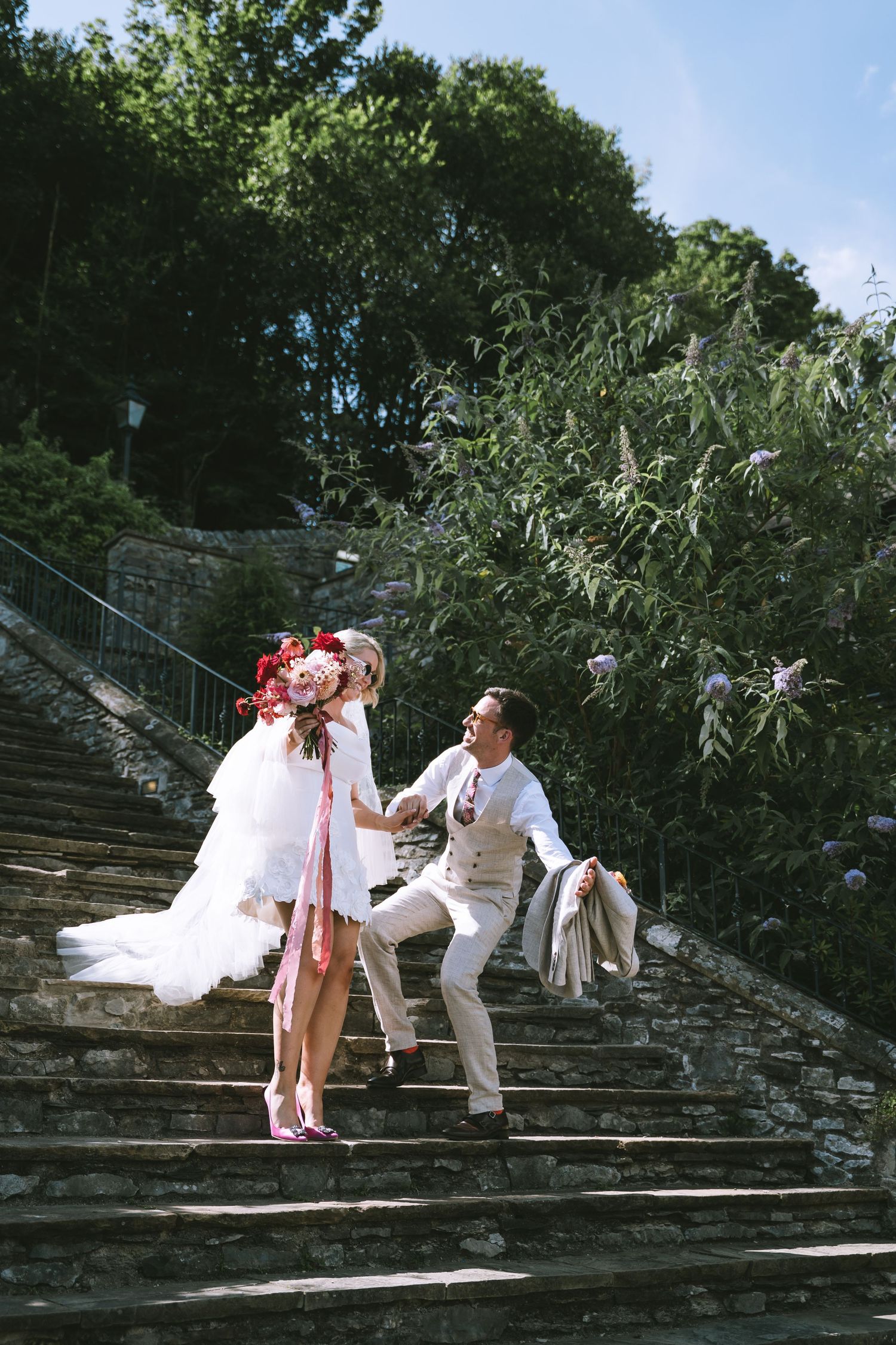 Happy bride and groom walk down stone steps outdoors, bride holding a stunning red and pink bouquet in Kendal, The Lake District