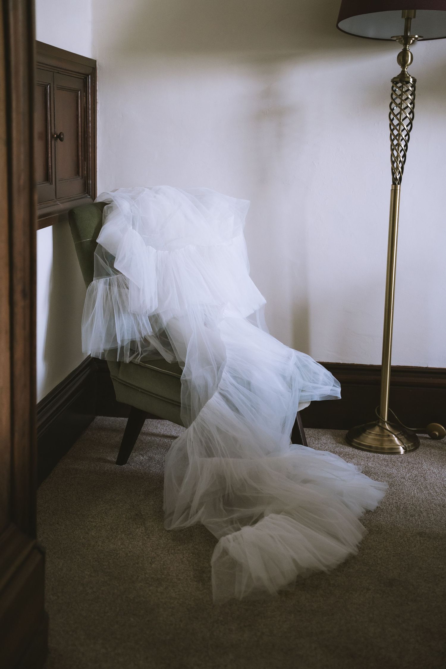 Elegant white tulle wedding veil draped over green armchair beside vintage brass floor lamp in hotel room.