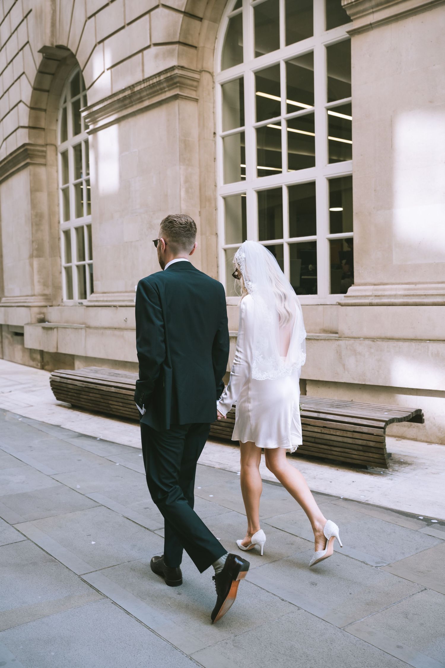 Bride in short white dress and veil walks hand in hand with groom in black suit along Manchester city street.