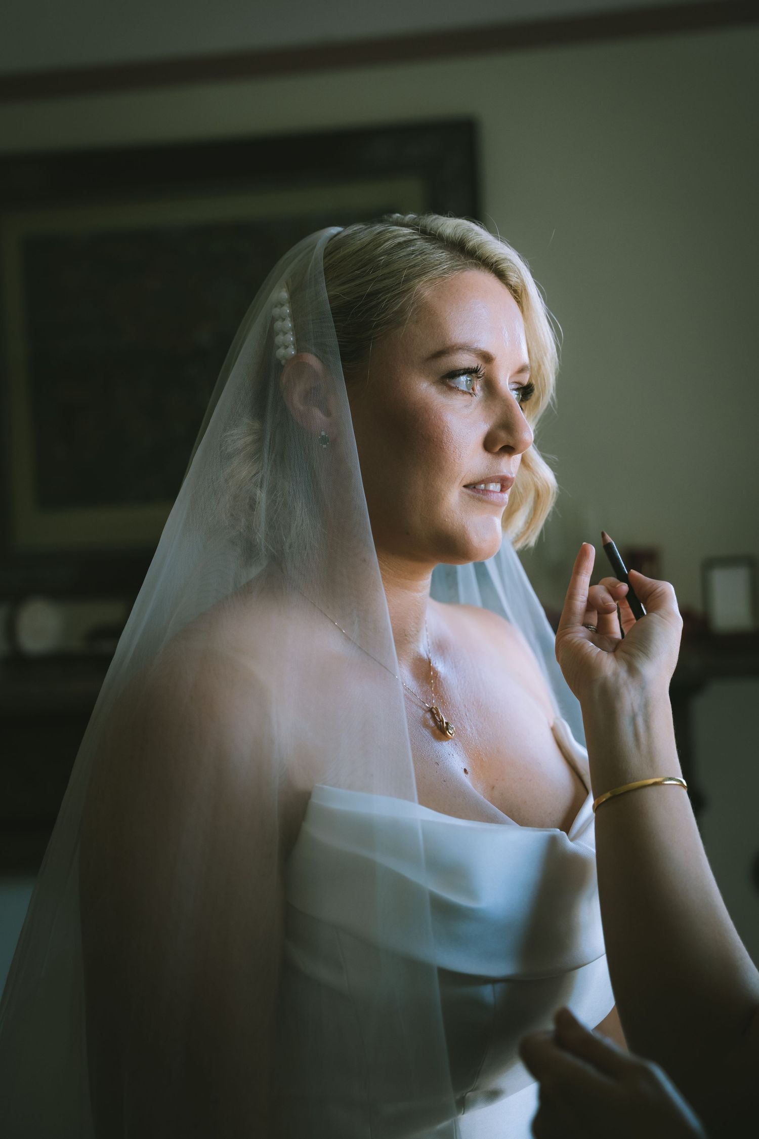 Beautiful blonde bride in a white strapless gown and veil gazes thoughtfully by a window on her wedding day in an AirBnb in Kendal