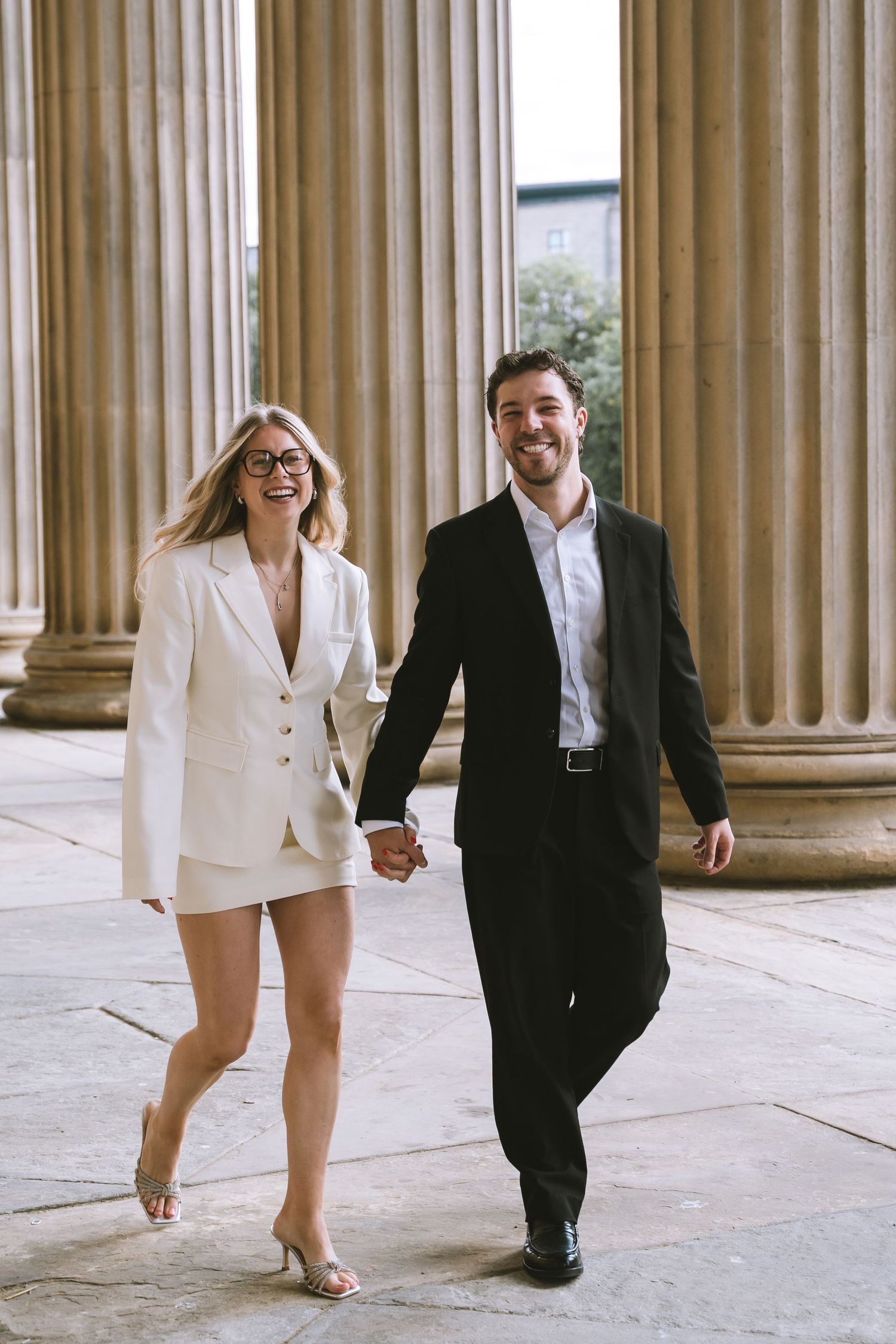 Smiling couple in wedding attire walk hand in hand between tall stone columns at a classical building in Liverpool at St Georges Hall