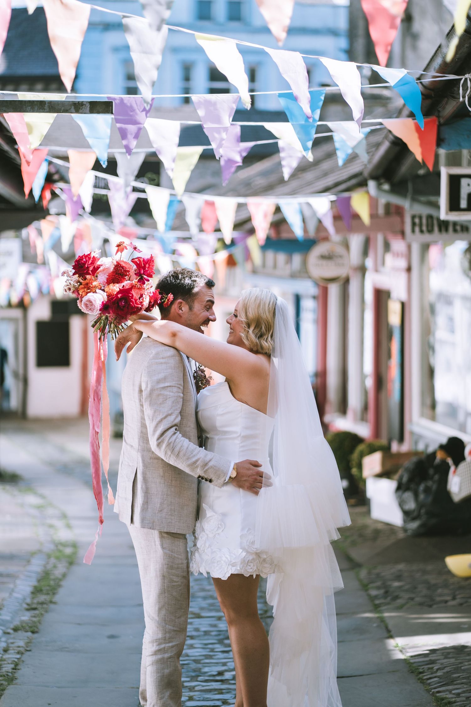 Bride and groom embrace on cobblestone street beneath colorful bunting with vibrant red bouquet in the streets of Kendal, The Lake District