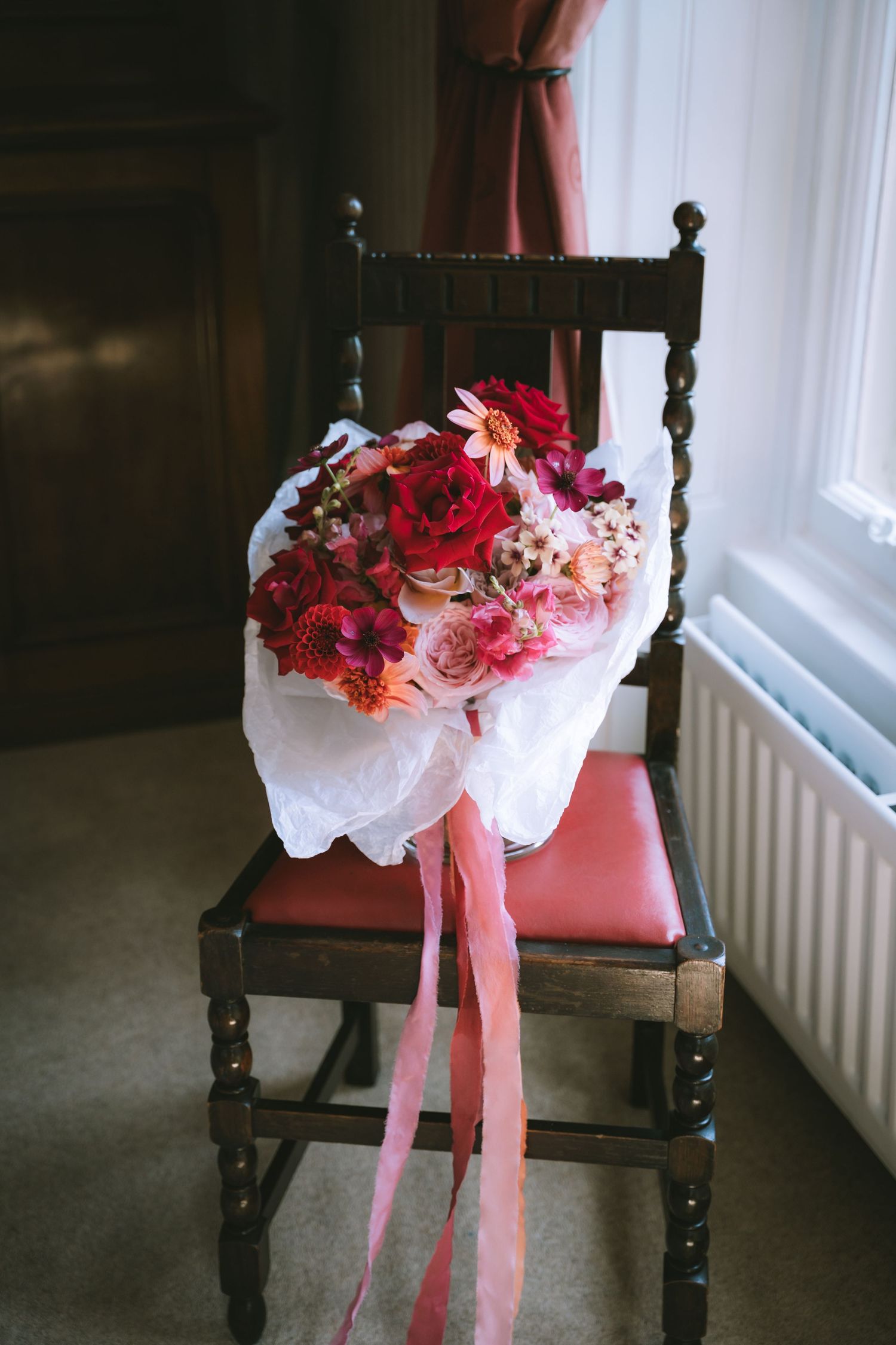 Elegant bridal bouquet of red and pink flowers wrapped in white tissue with pink ribbons resting on a wooden chair.
