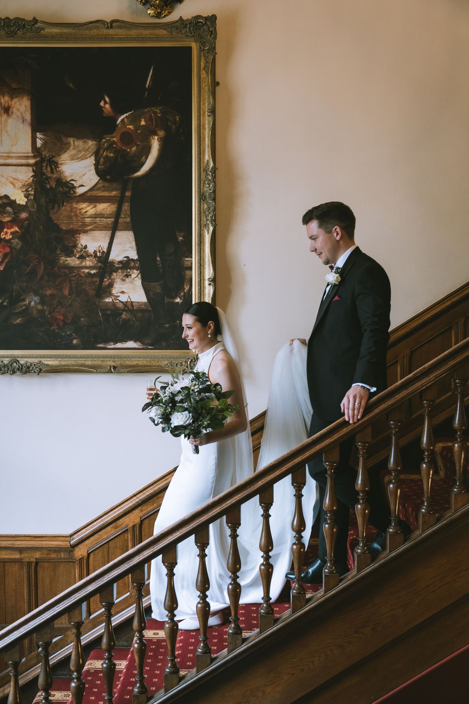 Bride and groom descend elegant wooden staircase in historic venue, with large oil painting on wall behind them at Walton Hall & Gardens