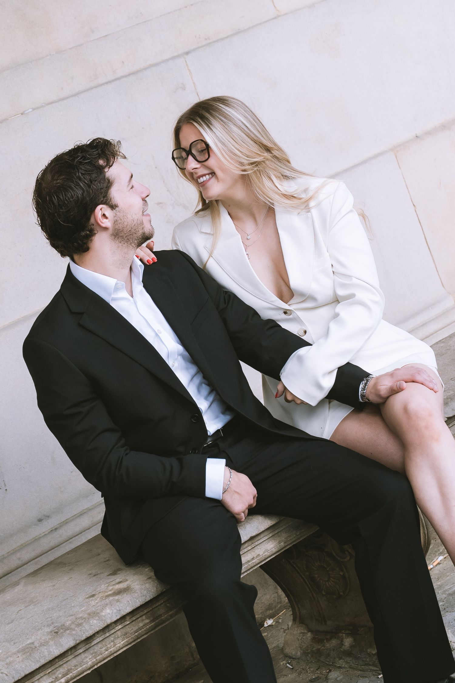 Bride in white blazer dress sits on stone bench smiling at groom in black suit at classical venue in Liverpool at St Georges Hall