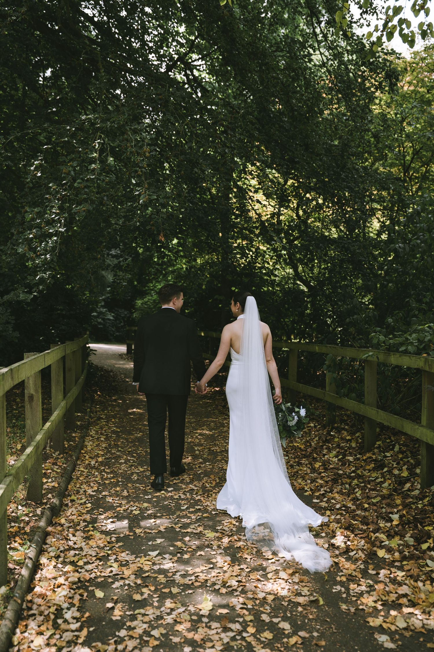 Bride and groom walk hand in hand across leaf-covered bridge through lush green woodland on their wedding day at Walton Hall & Gardens