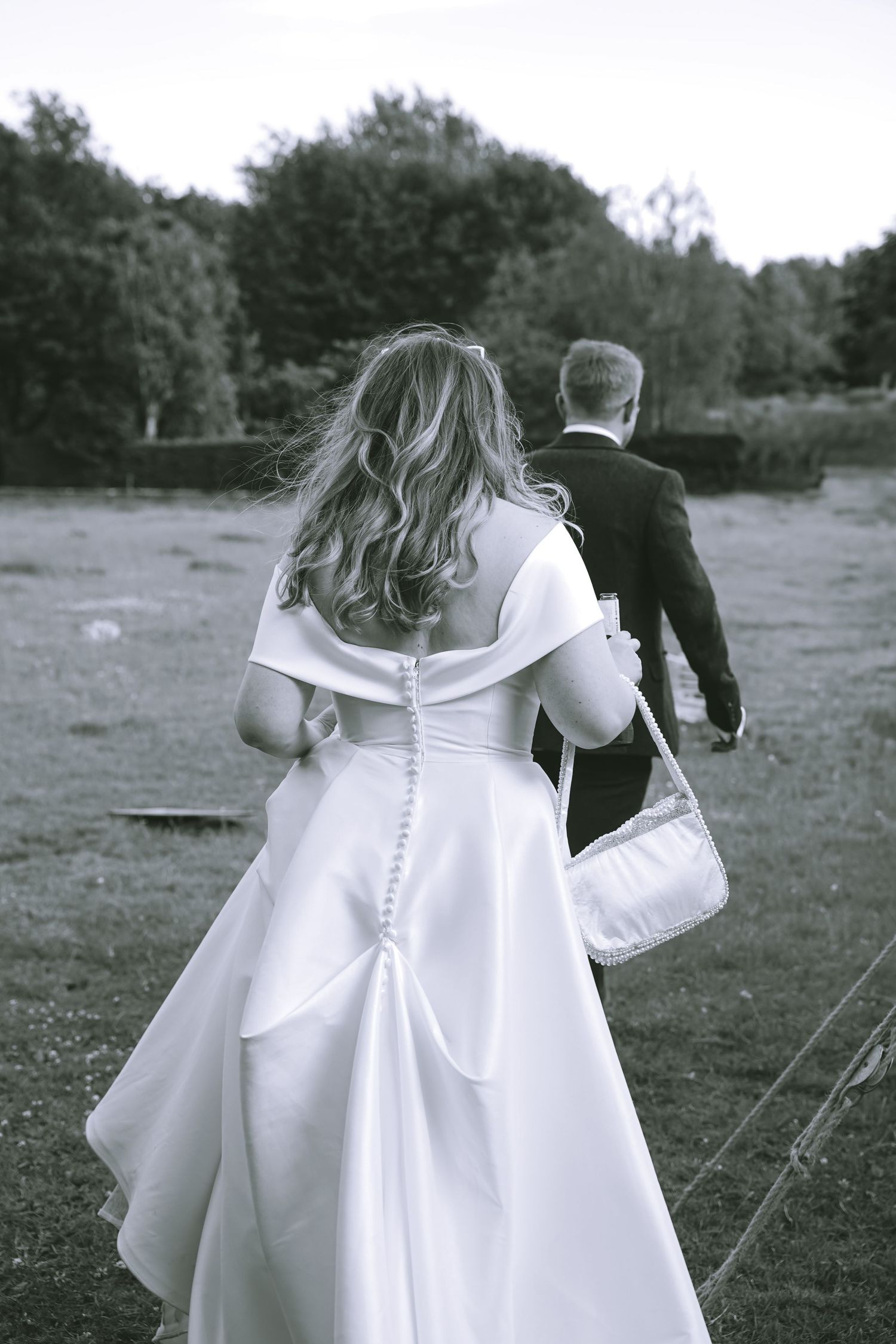 Black and white photo of a bride in a white gown with buttons walking beside a groom in a tuxedo outdoors.