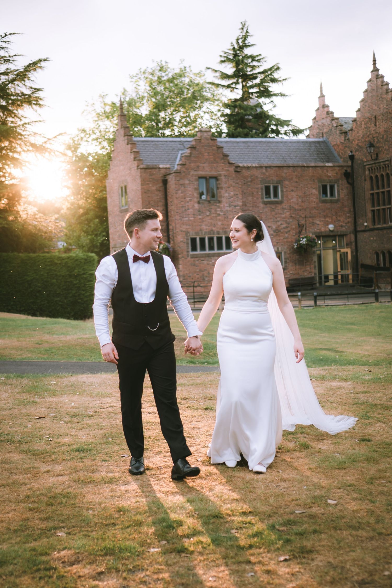 Bride and groom hold hands walking across lawn at golden sunset with historic brick manor house behind them at Walton Hall & Gardens
