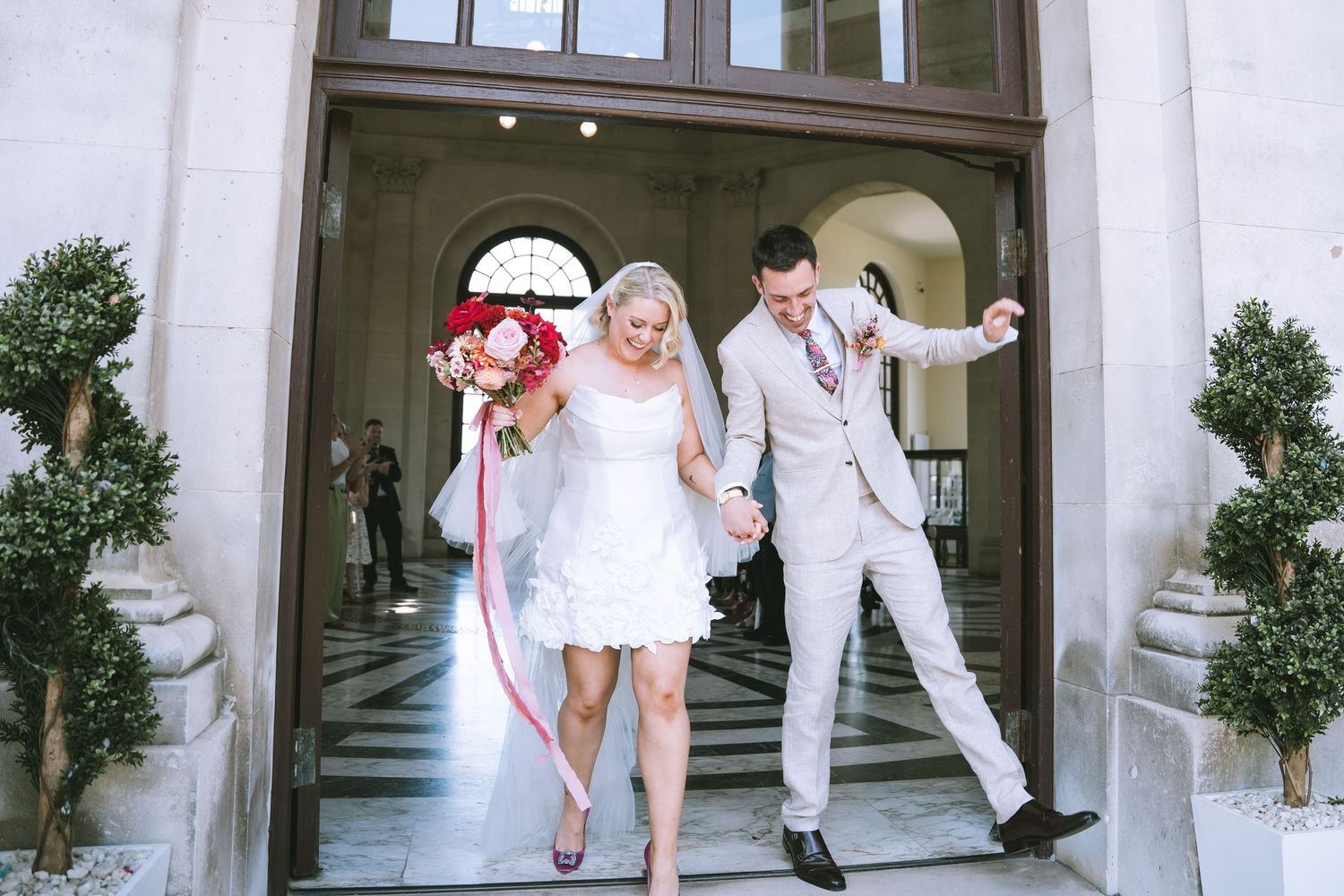 Joyful bride and groom exit a grand building, holding hands and carrying a vibrant red bridal bouquet in Lancaster at Ashton Memorial