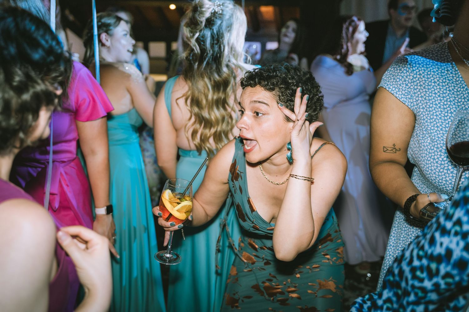 Woman in floral dress making a surprised expression while holding a cocktail on a crowded dance floor.