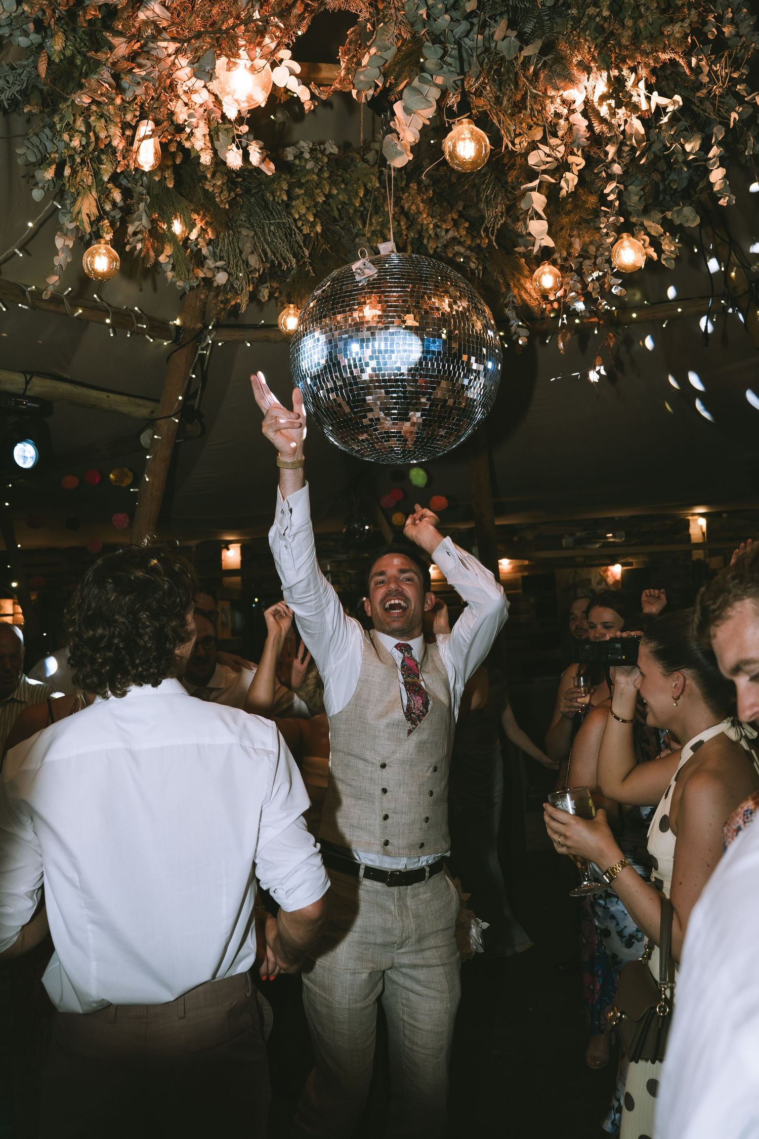 Joyful groom in vest raises hands under spinning disco ball surrounded by guests on wedding dance floor at Tipi Wedding at Kendal Brewery