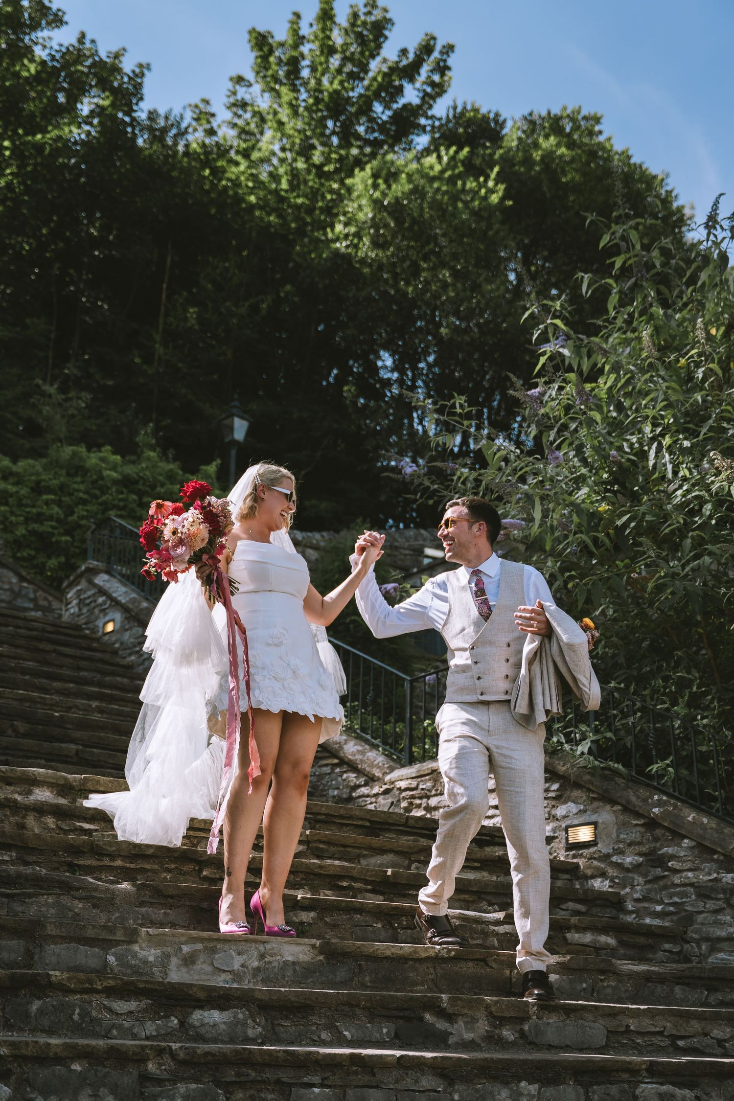 Bride and groom dancing joyfully on stone steps outdoors, surrounded by lush green trees on their wedding day.
