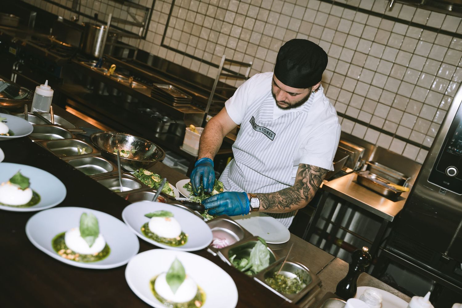 Tattooed chef in white uniform carefully plating elegant dishes in a professional restaurant kitchen.