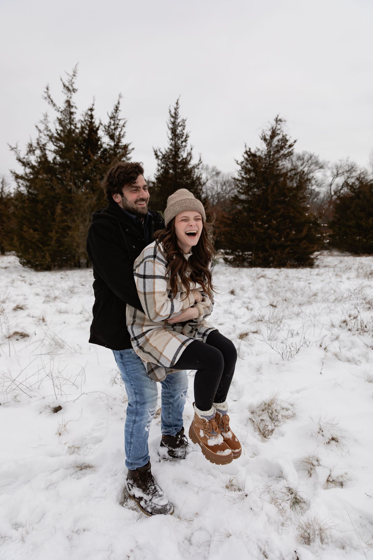 Two people playing and laughing together in a snowy winter landscape with evergreen trees in the background.