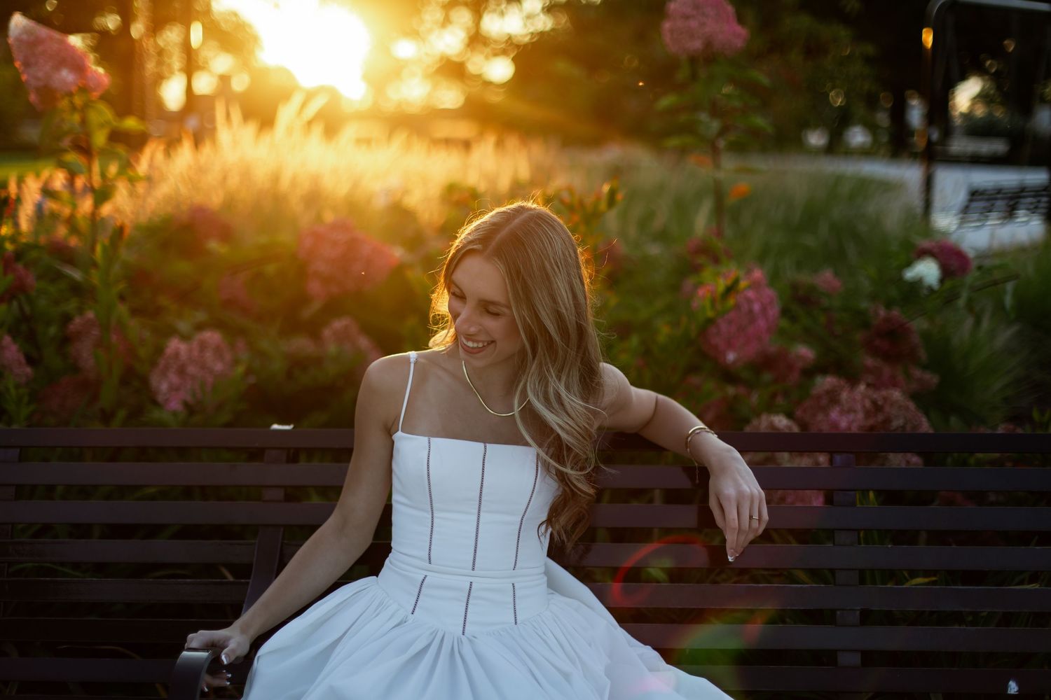 Bride in white wedding dress stands in garden at golden sunset with pink flowers in background.