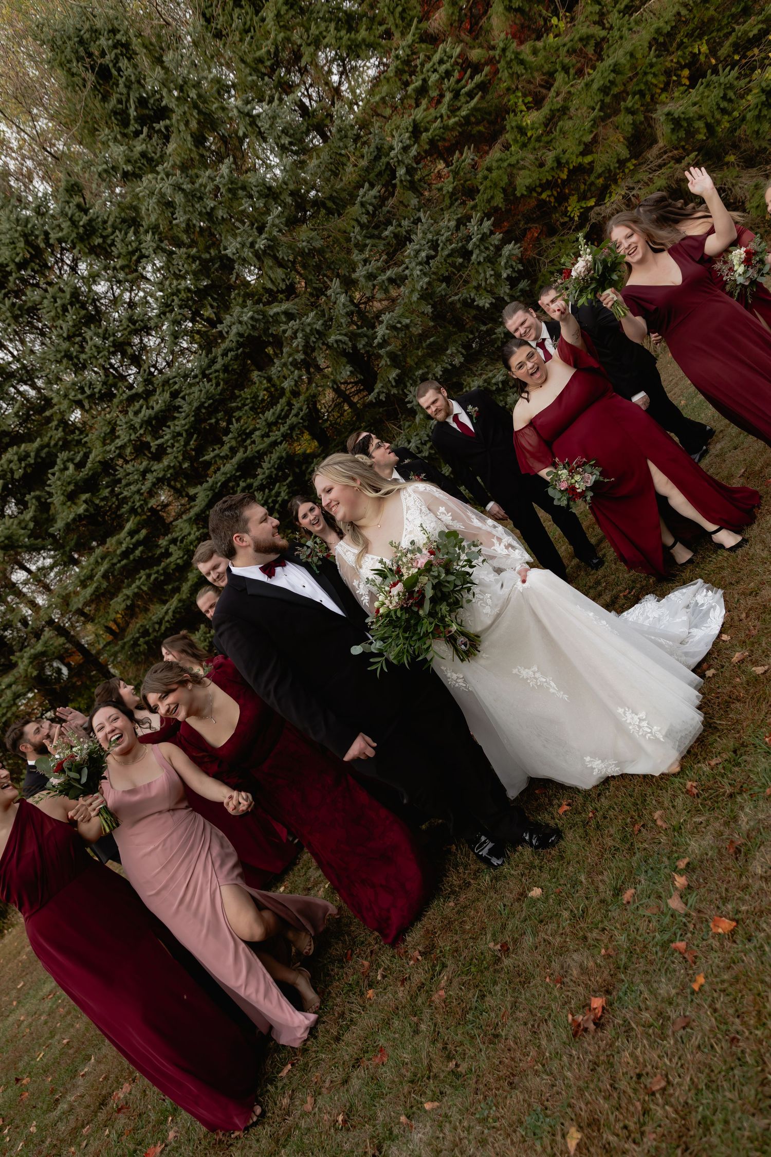 Wedding party poses for creative dip photos in burgundy and blush dresses against natural outdoor backdrop.
